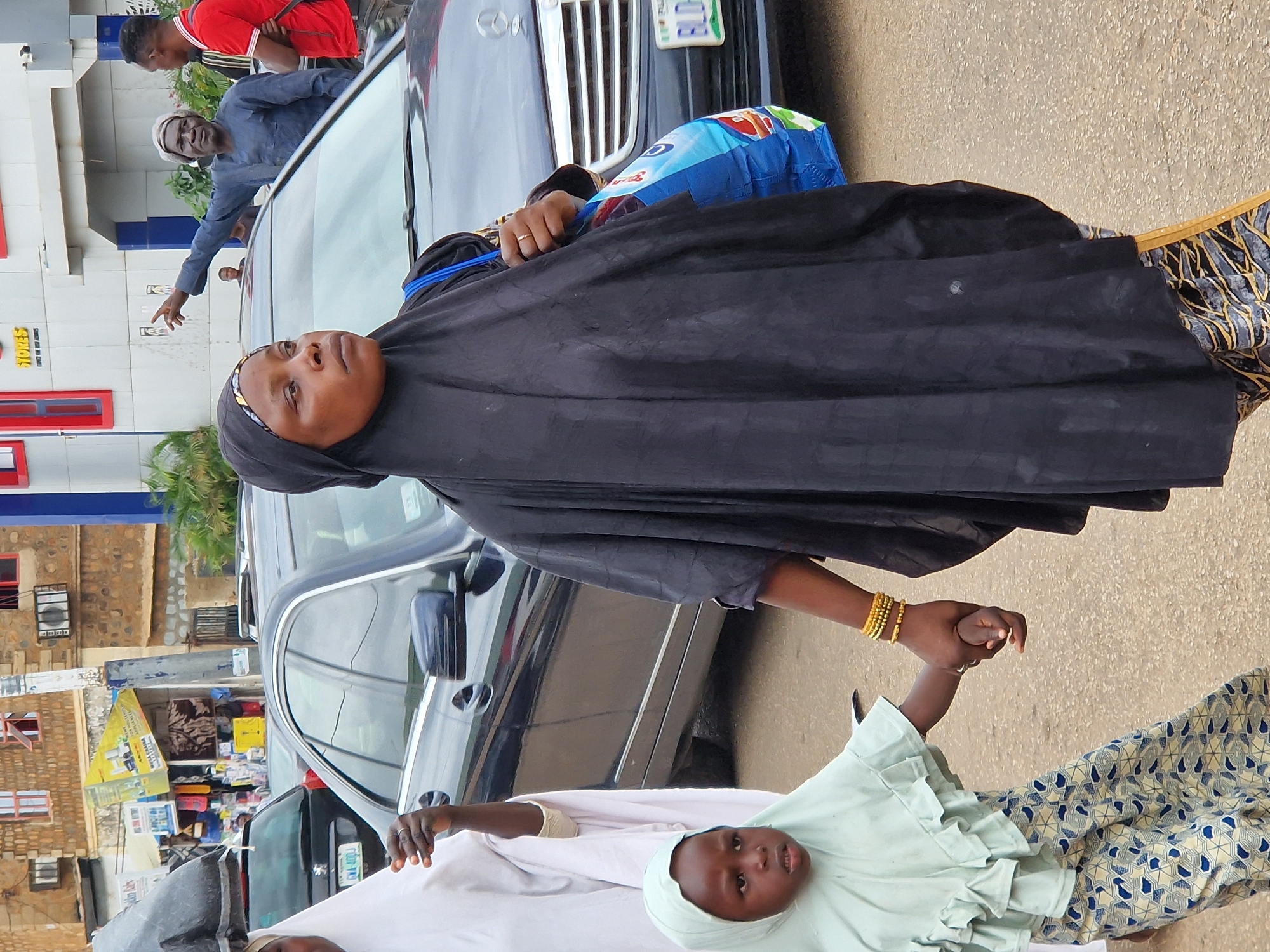 A vertically oriented, close-up shot shows a woman in a black hijab and dress holding the hand of a young child wearing a pale green headscarf and dress. The woman has a gold bracelet on her right wrist, and a ring on her left hand. The child's dress has ruffled sleeves. They are standing next to a dark-colored car. In the background, there is a building with stone walls and windows. There are some green plants visible. The lighting suggests it is daytime. A blue plastic bag is visible near the car's windshield. License plates are visible on the car, but the text is not legible.