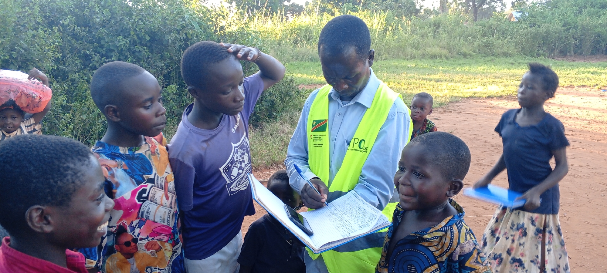 In Ngalula, Democratic Republic of the Congo, an adult male in a light blue shirt and a yellow safety vest stands outdoors, writing on a clipboard with a blue pen. He is surrounded by a group of children. Some children are looking at the clipboard, while others are looking elsewhere. A young child peers from behind a person carrying items on their head in the background. The environment includes trees and a dirt path. A few children appear to be holding folders.