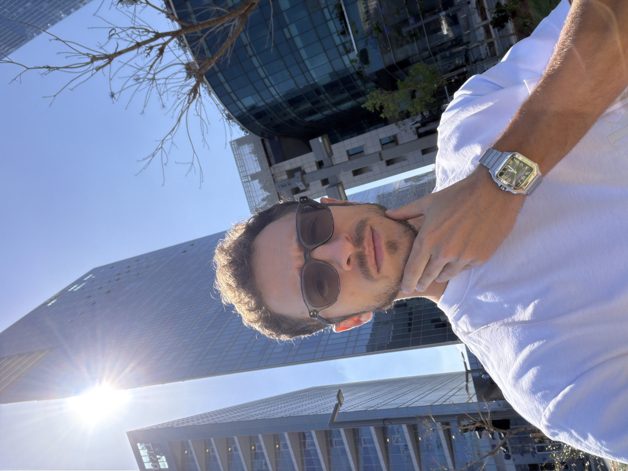 This is a rotated outdoor portrait of a man, likely a selfie, set against a backdrop of modern urban architecture and a clear blue sky. The man, with short dark hair and light stubble, wears dark sunglasses and a white t-shirt. His left arm is visible, featuring a silver-toned metallic watch with a green dial. The watch face clearly displays Roman numerals, the brand name "CARTIER" below the 12 o'clock position, and "AUTOMATIC" beneath the hands. His right hand is casually placed near his chin or neck.
The background consists of several contemporary glass skyscrapers with distinct geometric designs, one of which shows a prominent sunburst reflection, indicating bright daytime conditions with a clear sky. Bare tree branches are visible in the upper part of the frame, suggesting a specific season or type of urban landscaping. The modern buildings and bright sunlight suggest a vibrant urban environment, consistent with a city like Tel-Aviv, Israel. The overall impression is casual and bright.