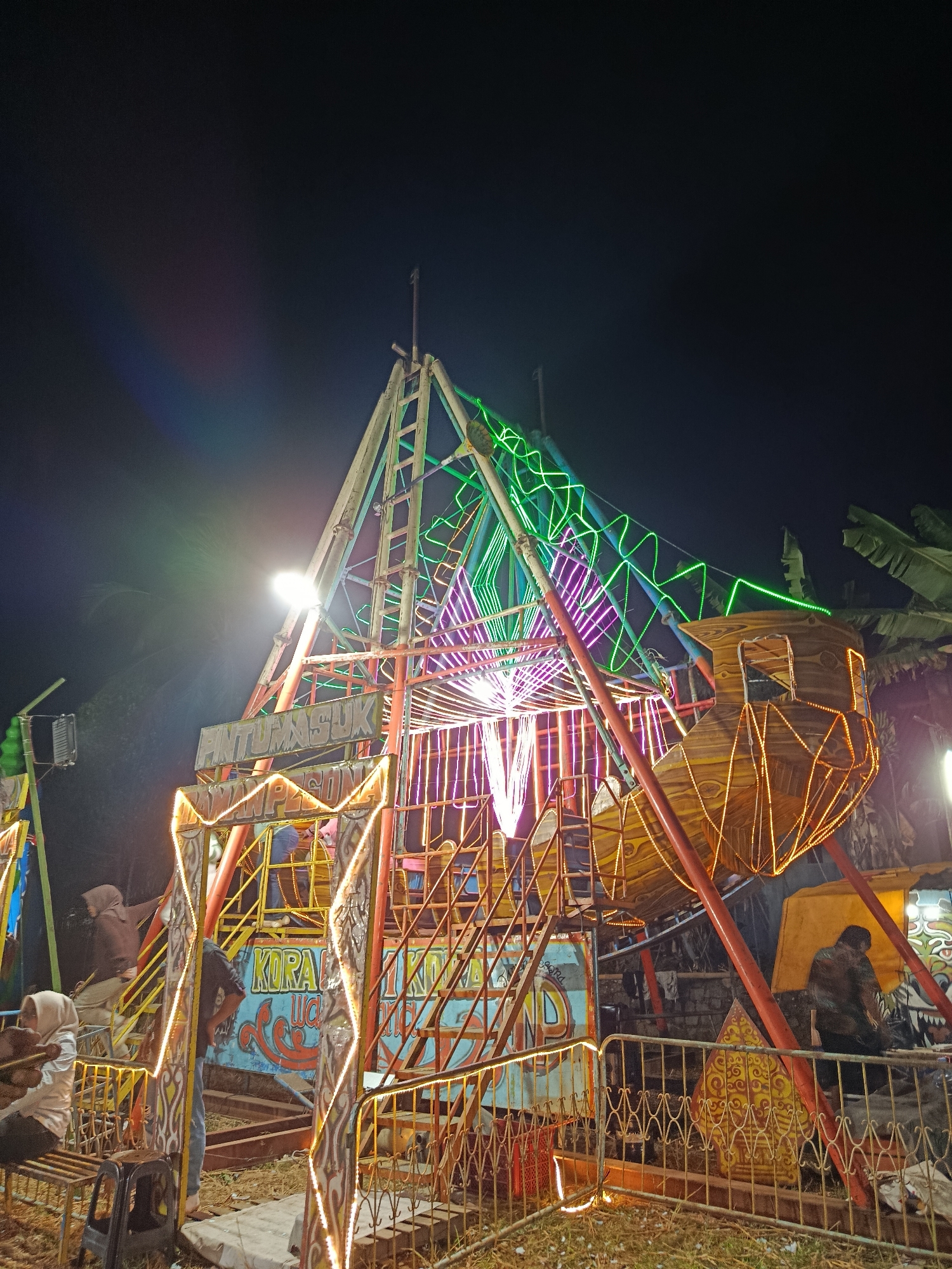 In the city of Sambirata, Indonesia, at night, a carnival ride is illuminated by a multitude of colorful lights. The ride, with a large central structure made of metal scaffolding, features a wooden boat-like gondola at its upper right. The structure is adorned with vibrant green and purple neon lights that trace its geometric design. Yellow and white lights outline the entrance and various platforms of the ride, casting a warm glow on the surrounding area.
Several individuals are visible around the ride. In the foreground, to the left, two people are seated on a bench, with one person wearing a light-colored headscarf and the other out of focus. To their right, a person in a dark shirt and blue jeans stands near the entrance. Further back, other individuals can be seen on the ride's platforms, some appearing to be preparing for or disembarking from the ride.
The ground beneath the ride is uneven, covered with dry grass and some scattered debris. A metal fence with a decorative pattern encloses part of the ride area. In the background, banana leaves are visible, suggesting a tropical environment. The overall scene captures the lively atmosphere of a night-time fairground.