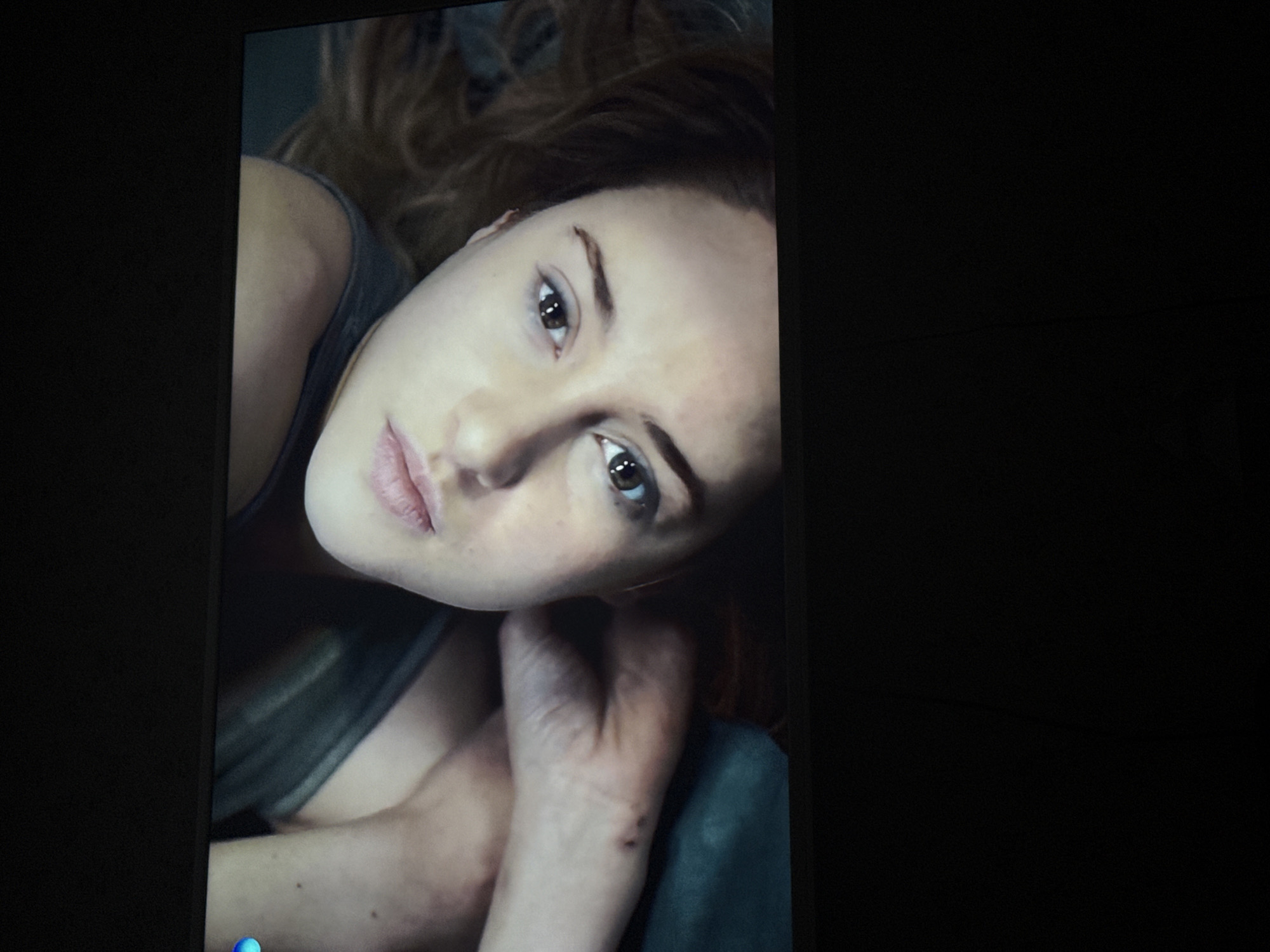 A close-up, tilted shot captures a woman with brown hair and brown eyes. She is wearing a gray tank top. Her head is tilted towards her hand. There are small marks on her hand and forearm. The background is predominantly dark, with a black surface to the right and blurry elements to the left. No location data from Mytishchi, Russia, is discernible from the image.