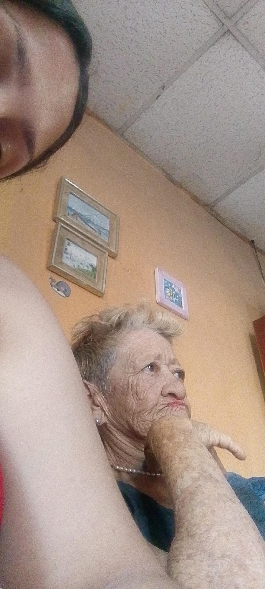 In this media file, an elderly woman is captured in a close-up, profile shot. She has light-colored, short, permed hair and her face is deeply lined, indicative of her age. Her eyes are open, and she appears to be looking off to the right of the frame. Her hand is raised, with her fingers touching her chin, suggesting a pensive or thoughtful mood. She is wearing a string of pearls around her neck and an earring in her visible ear. The background consists of an orange-colored wall with two framed pictures hung on it. One picture appears to be a seascape or landscape, and the other is smaller and lighter colored, possibly with a floral design. A small decal of a whale is visible on the wall below the frames. To the left of the woman, a portion of another person's body, likely their arm and shoulder, is visible, suggesting they are in close proximity and possibly taking the photo. The ceiling in the upper portion of the image is made of acoustic tiles, commonly found in interior spaces. The lighting suggests an indoor setting, likely during daylight hours given the overall brightness, though no specific time of day can be definitively determined. The context provided indicates the location as Juan Griego, Venezuela.