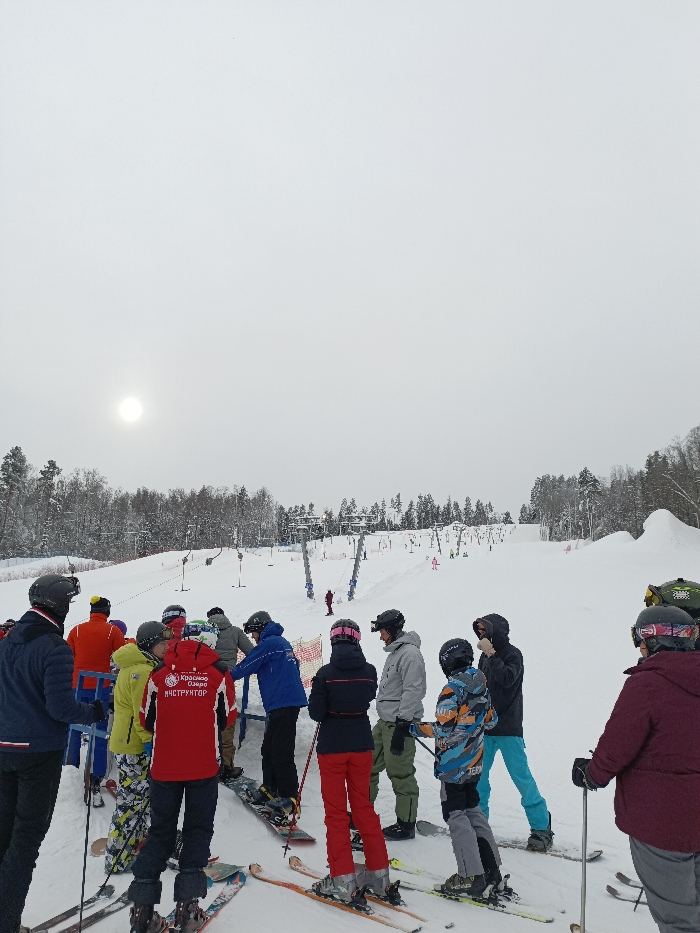 The image depicts an active ski resort environment at Krasnoye Ozero, located near Svetloe, Russia. Approximately 15 individuals, dressed in various brightly colored winter sports attire, helmets, and goggles, are gathered on a snow-covered slope in the foreground, predominantly at what appears to be the base of a ski lift queue. Most individuals are on skis, with at least one person equipped with a snowboard. A prominent figure in a red jacket, facing away from the viewer, displays the text "КРАСНОЕ ОЗЕРО ИНСТРУКТОР" (Krasnoye Ozero Instructor) on the back. A yellow and orange mesh barrier is partially visible behind some of the individuals.
The midground features an ascending snow-covered ski slope equipped with multiple T-bar or platter-style ski lifts, their poles extending upwards. Several other skiers and snowboarders are visible on the slope and near the lift lines. The background consists of a dense, snow-laden coniferous forest bordering the ski runs, under an overcast sky with a dimly visible, hazy sun disk in the upper left quadrant. Sculpted snow features are observable on the far right midground. The overall scene indicates a winter day with diffused natural light.