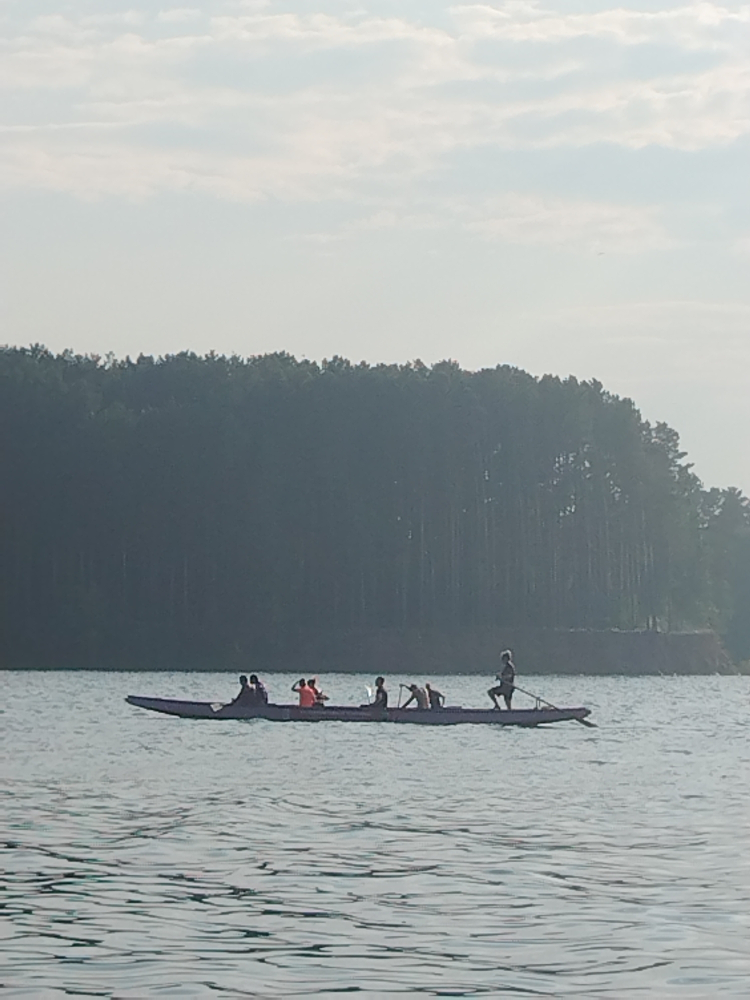 A long, narrow boat, likely a canoe or kayak, glides across calm, rippling water. Silhouetted against the hazy sky and a dense, dark green forest line, several people are visible within the boat. Some appear to be paddling, while others are seated. One person at the stern of the boat is standing and holding an oar or paddle, seemingly steering. The overall scene suggests a peaceful outdoor activity, possibly recreational canoeing or a similar water sport. The time of day appears to be either early morning or late afternoon, given the soft, diffused light and the hazy atmosphere. The background shows a prominent treeline, indicative of a natural setting, possibly a lake or a wide river. The weather seems mild, with a light overcast sky. The location cues, such as the forest and water, are consistent with a natural environment. No text is visible in the image.