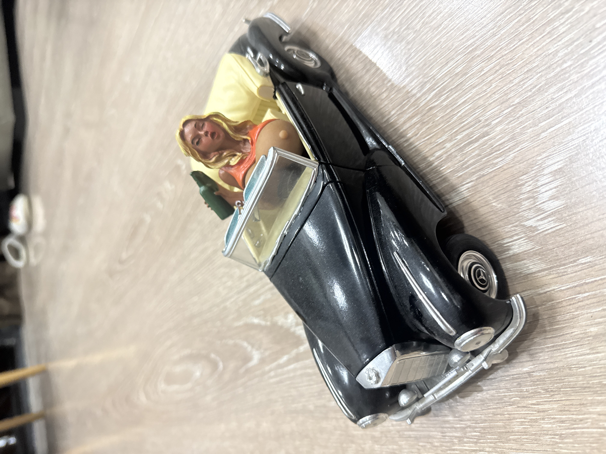 A high-angle, medium shot captures a black 1950s-style convertible toy car on a light-colored wooden floor. The car's interior is lined with yellow fabric. A blonde, female figurine with an orange top is seated in the passenger seat, holding a green bottle. The figurine's chest is exposed. The background is blurred, showing parts of furniture and out-of-focus objects, suggesting an indoor environment. There is no information to suggest this scene is located in Tbilisi, Georgia.