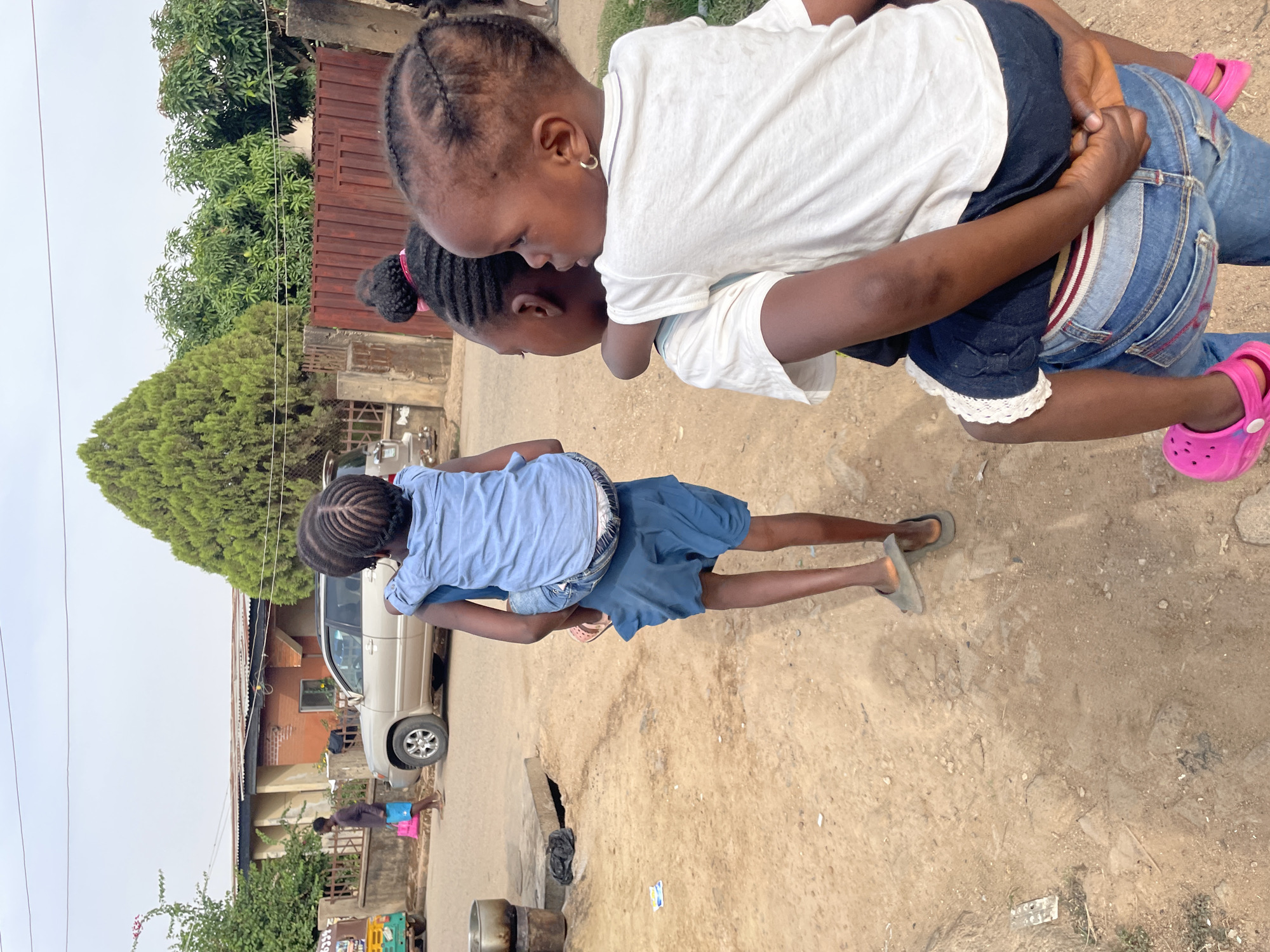 In a city street in Jos, Nigeria, one child carries another on their back. The child on the bottom wears a blue outfit and gray sandals. The child on top wears a white shirt, denim jeans, and pink sandals. Trees and a brown metal fence are in the background. The street surface is dirt. A silver vehicle and a building are visible along the road.