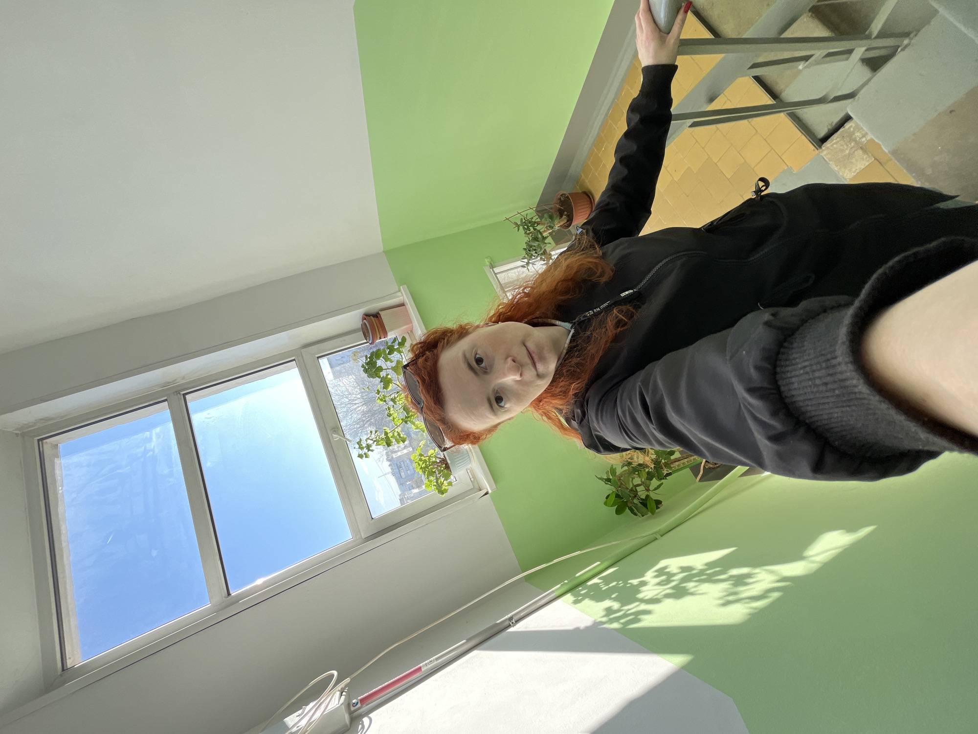 A woman with red hair stands in a stairwell painted white and light green. She wears a black jacket. A window provides a view of the sky and buildings, potentially in Chaikovsky, Russia, though specific landmarks are not visible. Potted plants are near the window and along the green wall. The stairwell has metal railings and a yellow tiled floor on the upper levels. The photo seems to be a selfie taken from a low angle.
