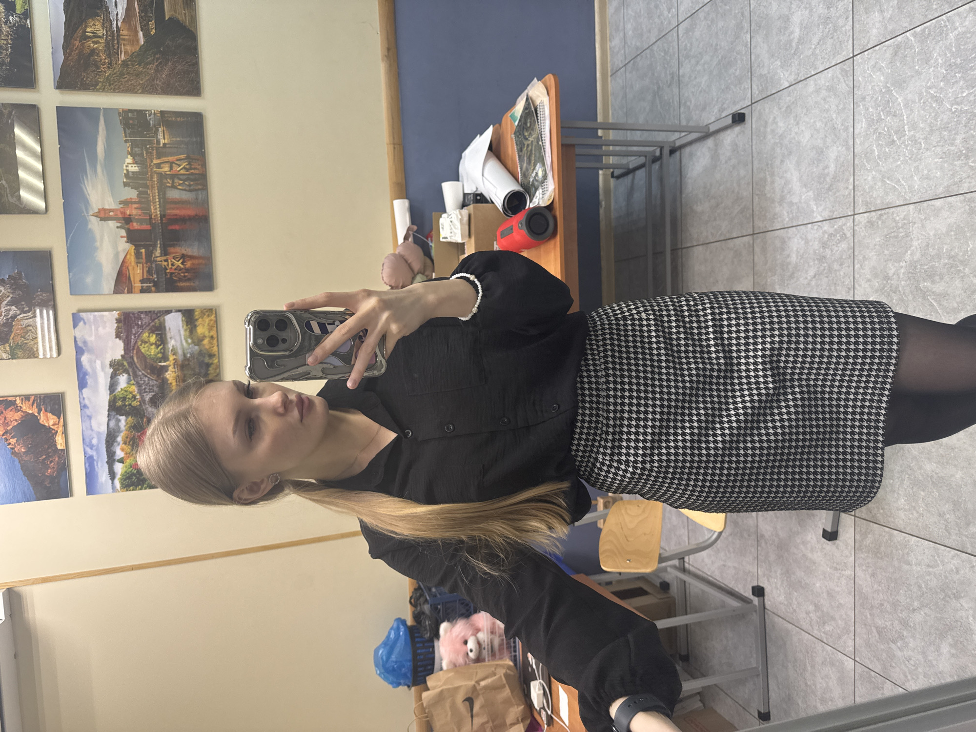 A fair-skinned female is posing for a selfie in a classroom. She is wearing a black button-down shirt, a houndstooth skirt, and dark tights. Her long blonde hair is pulled back in a ponytail. She is holding a smartphone in her left hand, and her right hand is adorned with a beaded bracelet.
Behind her, a wall is decorated with landscape photographs. To the right, a desk is cluttered with papers, containers, and a red speaker. Further in the background, there are additional desks and chairs, indicating a classroom setting.
Given the presence of landscape photographs and other details, there is no forensic information to indicate this image was taken in Perm, Russia.