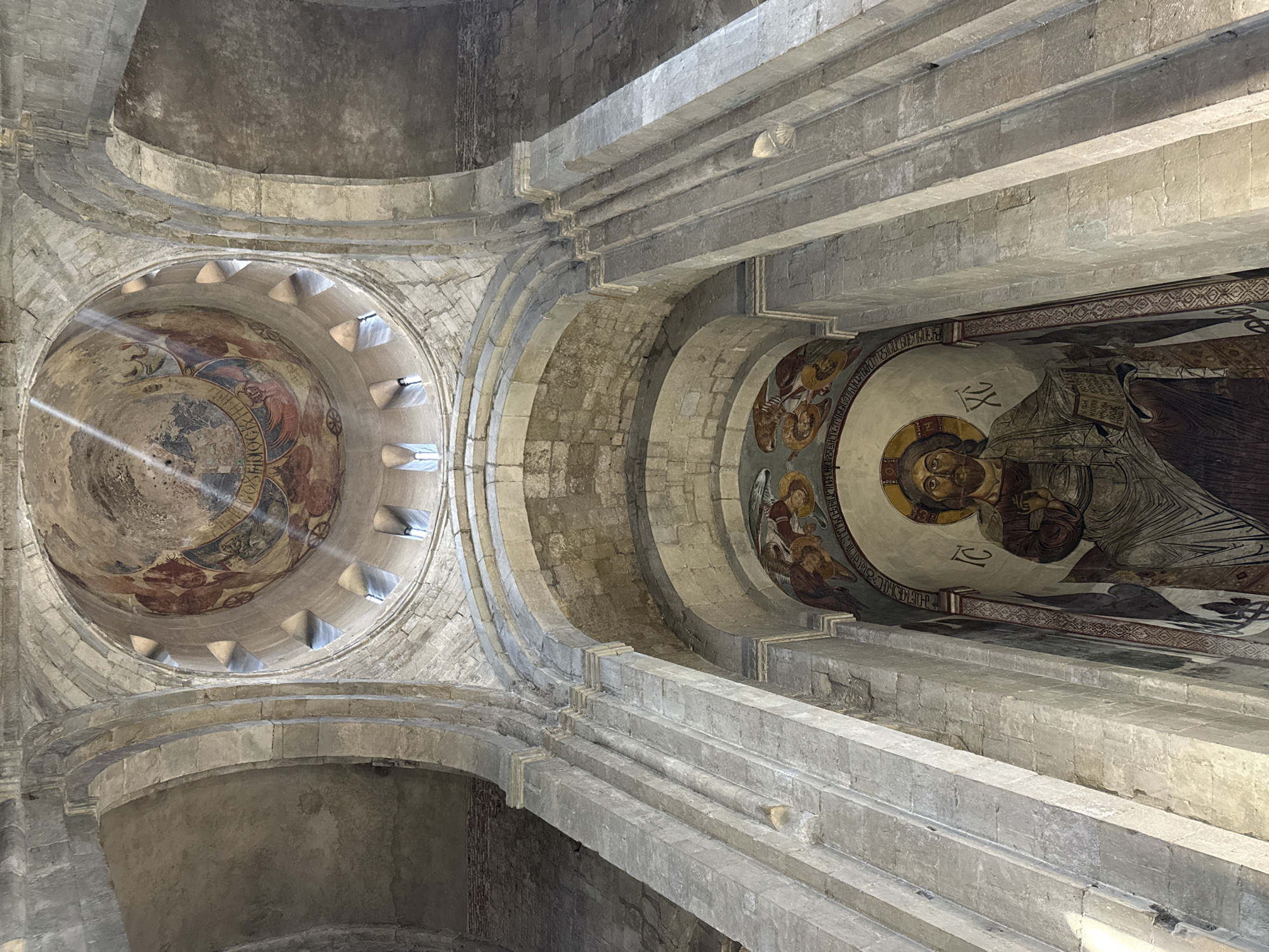 The image depicts the interior of the Svetitskhoveli Cathedral in Mtskheta, Georgia, looking up towards the dome and an apse fresco. Sunlight streams through arched windows in the dome, illuminating parts of the ancient frescoes and stone architecture. The dome features religious imagery with figures and circular designs. Below, within a large arched niche, is a fresco of Christ Pantocrator, with a halo and beard, holding a book. Surrounding Christ are angelic figures and inscriptions. The architecture is characterized by massive stone blocks and arched structures, indicative of medieval construction. The overall scene conveys a sense of history, spirituality, and architectural grandeur.