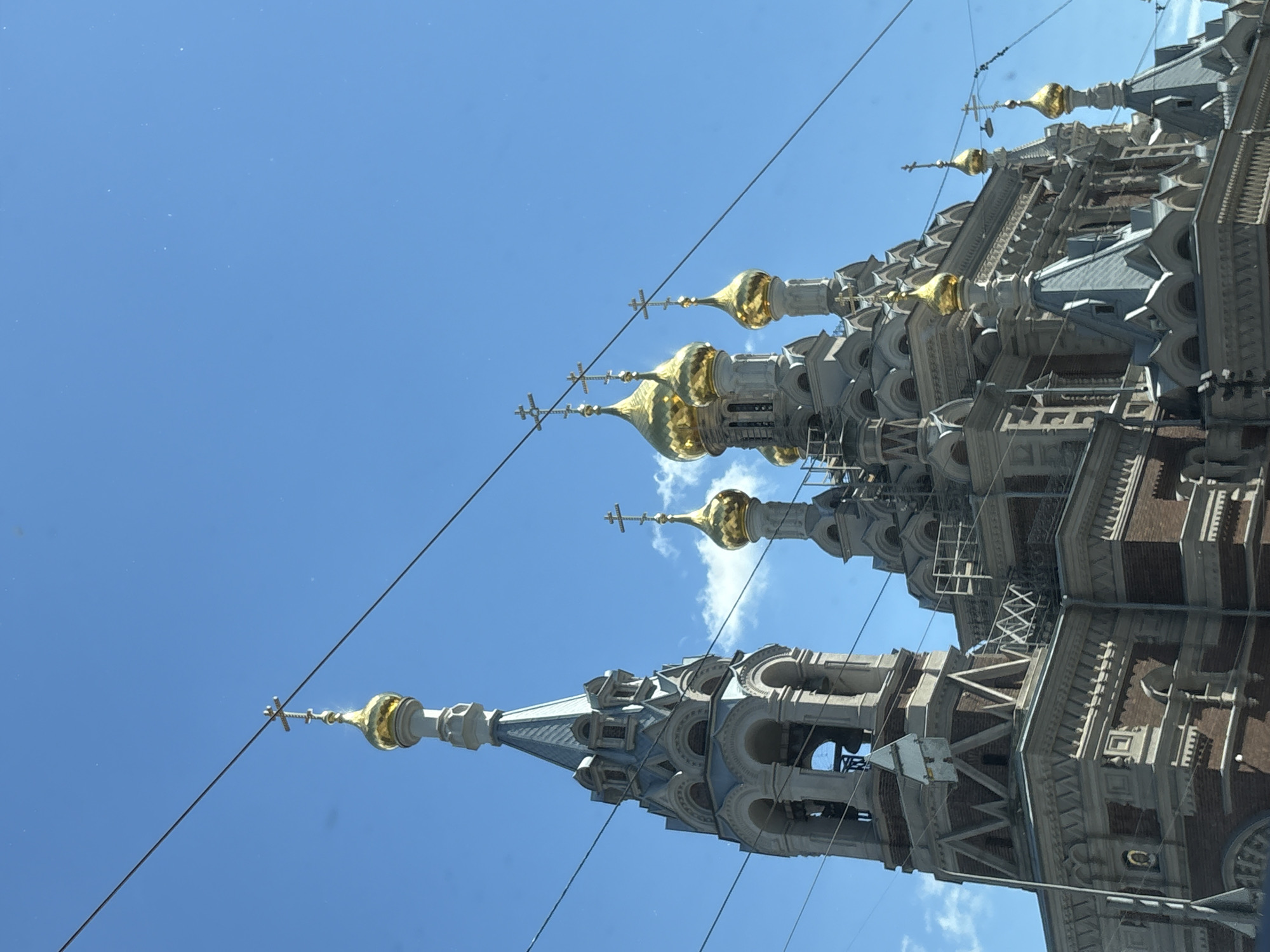 The image shows a close-up, low-angle view of the Alexander Nevsky Cathedral in St. Petersburg, Russia, on a clear day with a bright blue sky. The cathedral features multiple golden domes with crosses and intricate architectural details. The foreground is dominated by the cathedral's towers and domes, with some wires visible in front of the structure, suggesting the photo may have been taken from a moving vehicle or across a street. The lower portion of the cathedral is made of red brick, while the upper sections are adorned with white and grey decorative elements. The overall scene conveys a sense of grandeur and religious significance.