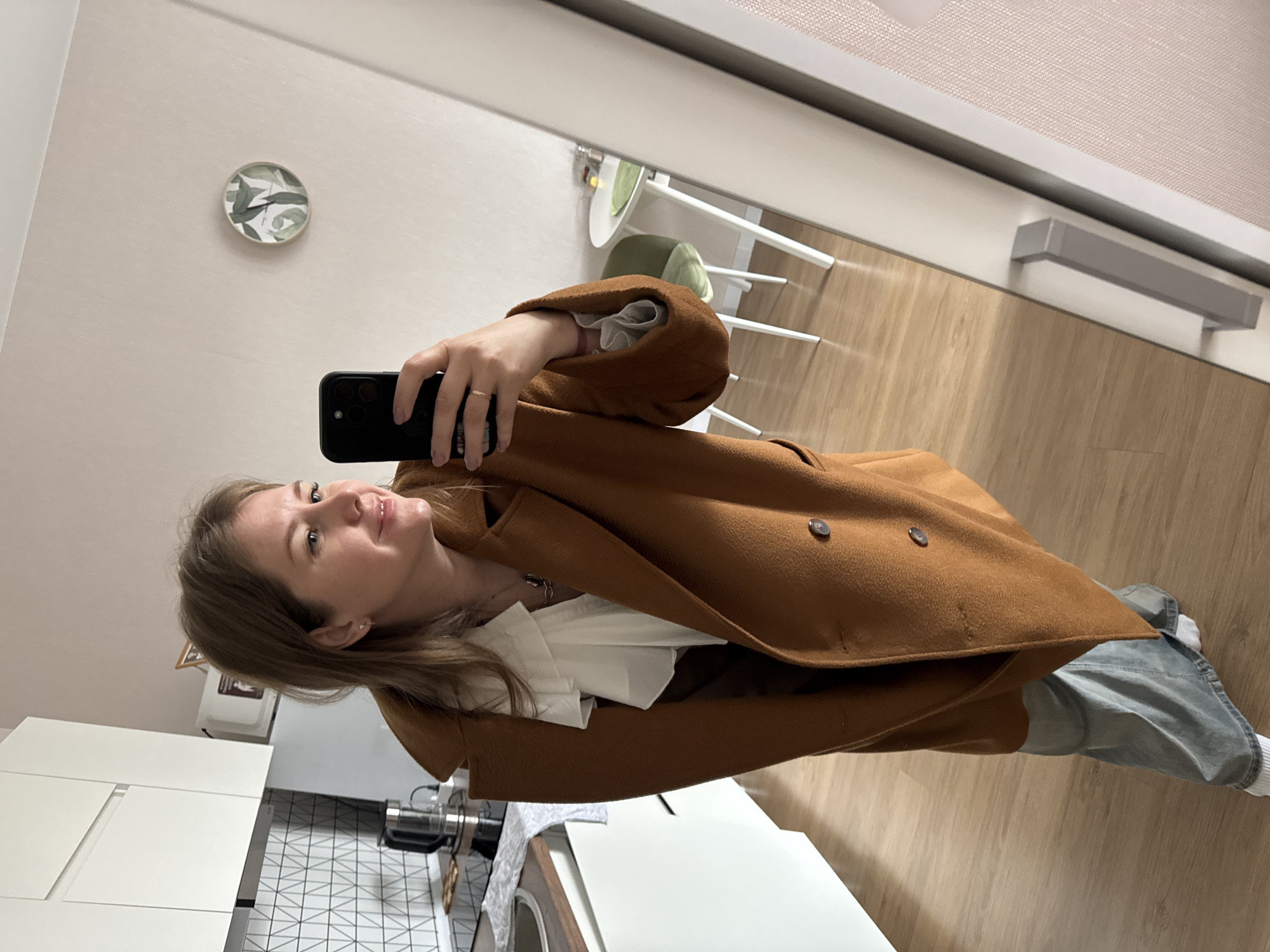 A woman is depicted taking a mirror selfie, appearing to be in an apartment or home setting. She has light brown hair, a fair complexion, and is smiling with a contented expression. She is wearing a warm-looking, brown or rust-colored overcoat, a white ruffled blouse, light blue jeans, and white socks. On her right hand, she wears a small ring on her ring finger, and she holds a black smartphone to take the picture. A small stud earring is visible in her right ear.
The background, which is a reflection in the mirror, shows a brightly lit room with light-colored walls and a light wood floor. A small, round piece of wall decor featuring green leaf patterns is visible. There's also a white table with a green cushion on a chair, and what appears to be window blinds or curtains reflected above.
In the actual foreground, to the left and below the woman, is a modern kitchen area. It features white cabinets, a black and white geometric patterned backsplash, a stainless steel sink, and what looks like a coffee machine or similar appliance on the countertop. The lighting suggests it is daytime. The overall aesthetic is clean and contemporary. No discernible text is visible in the image. Given the location context of Perm, Russia, this is likely a contemporary apartment interior there.