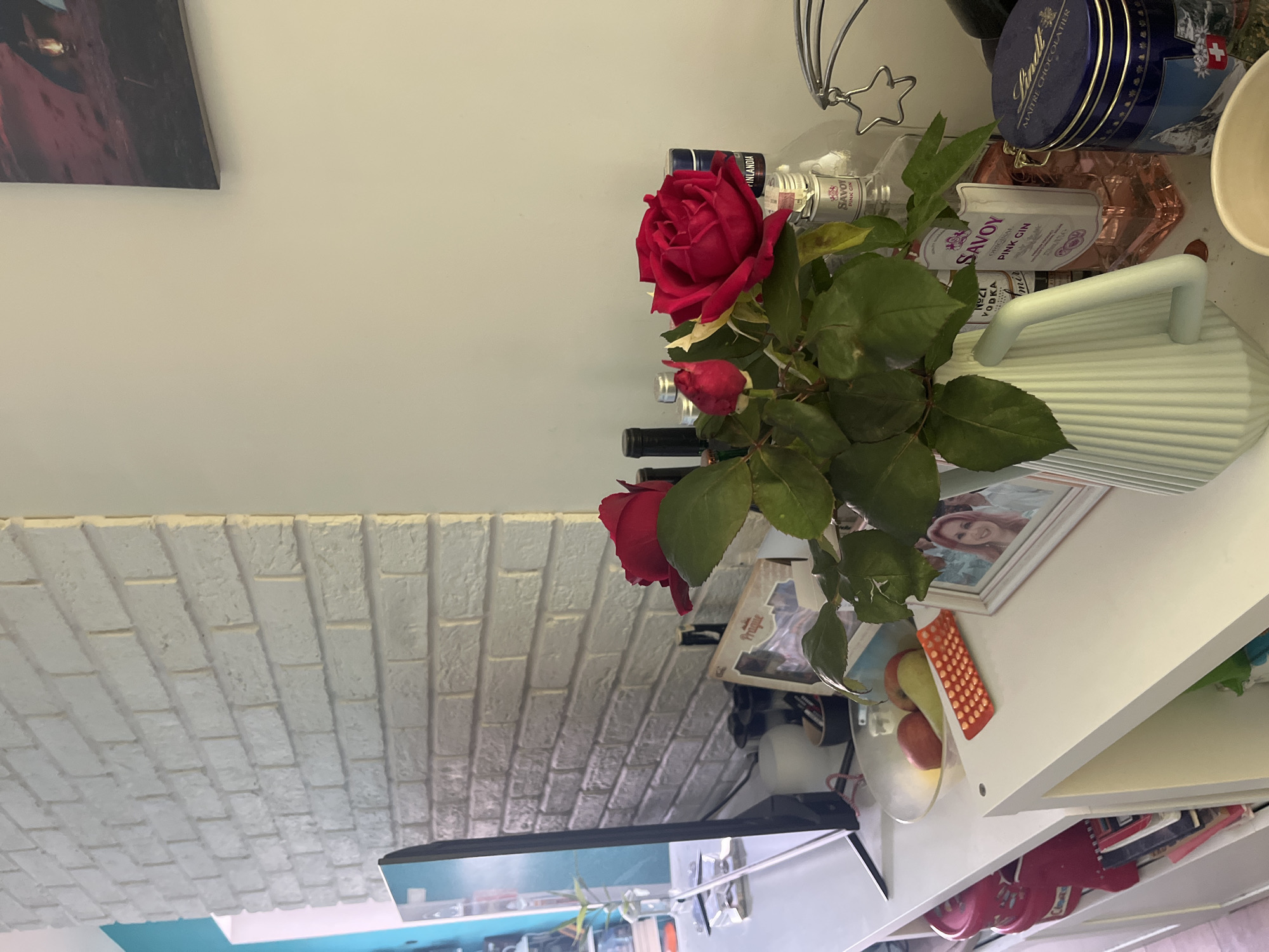 A still-life composition features a vase of red roses and greenery placed atop a white shelf. The shelf also holds a variety of items, including bottles of alcohol, a blue tin, a bowl with fruit, framed photos, and other miscellaneous objects. The background includes a white wall with a piece of artwork and a textured wall that mimics the appearance of white bricks. The composition is set in a room with a modern and clean aesthetic. The location is not identifiable as Pokrovnik, Bulgaria from the image alone.