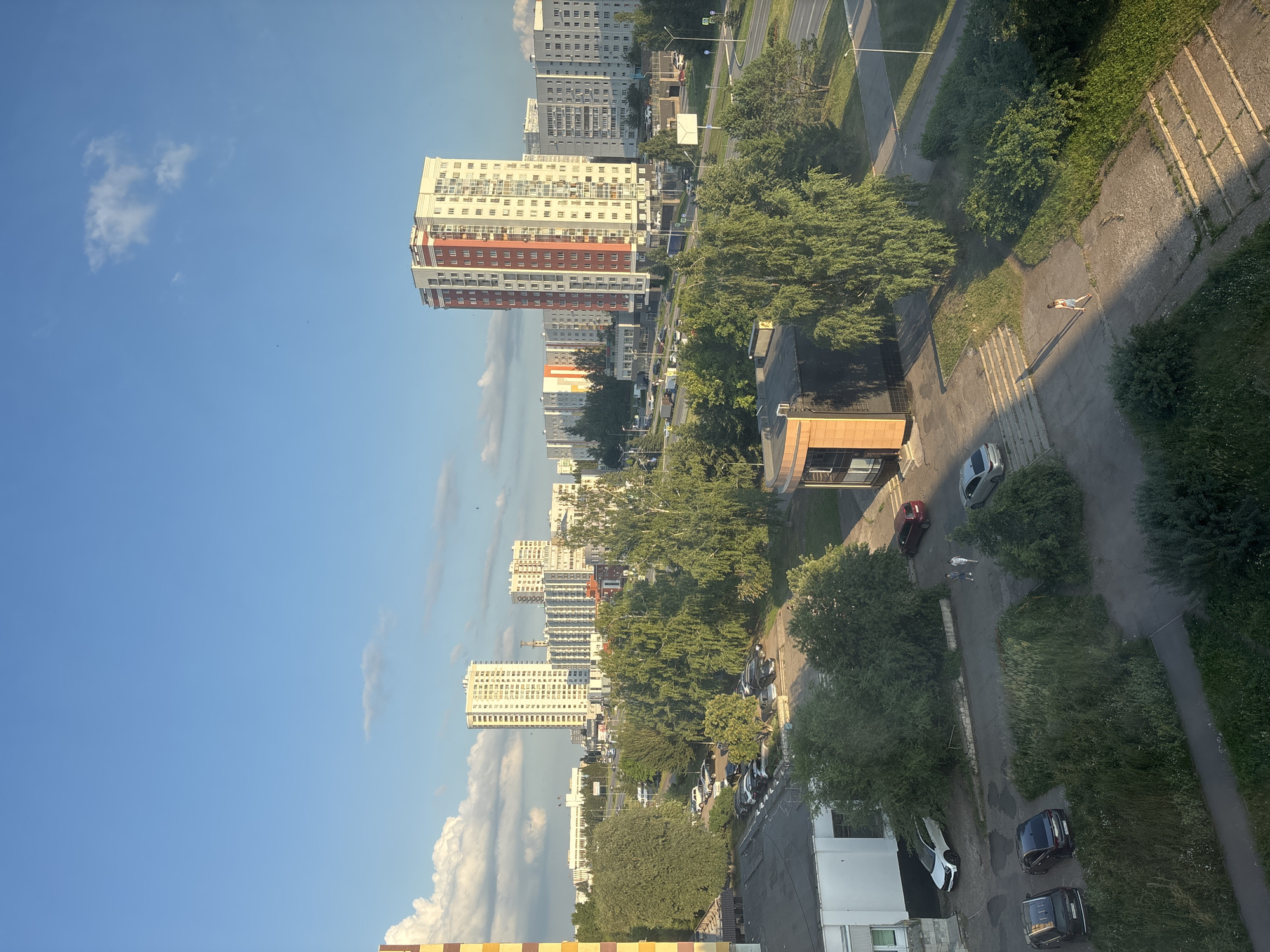 The image depicts an urban scene in Naberezhnye Chelny, Russia, under a clear blue sky with scattered clouds. In the foreground, there are several cars parked in a paved area adjacent to a low-rise building with white walls and a dark roof. A few people are walking in the paved area. Further in the background, numerous trees line a road with more parked cars and traffic. Dominating the mid-ground and background are tall, multi-story residential buildings. One prominent building features a red and white facade. The overall environment suggests a residential district with green spaces and typical city infrastructure.
