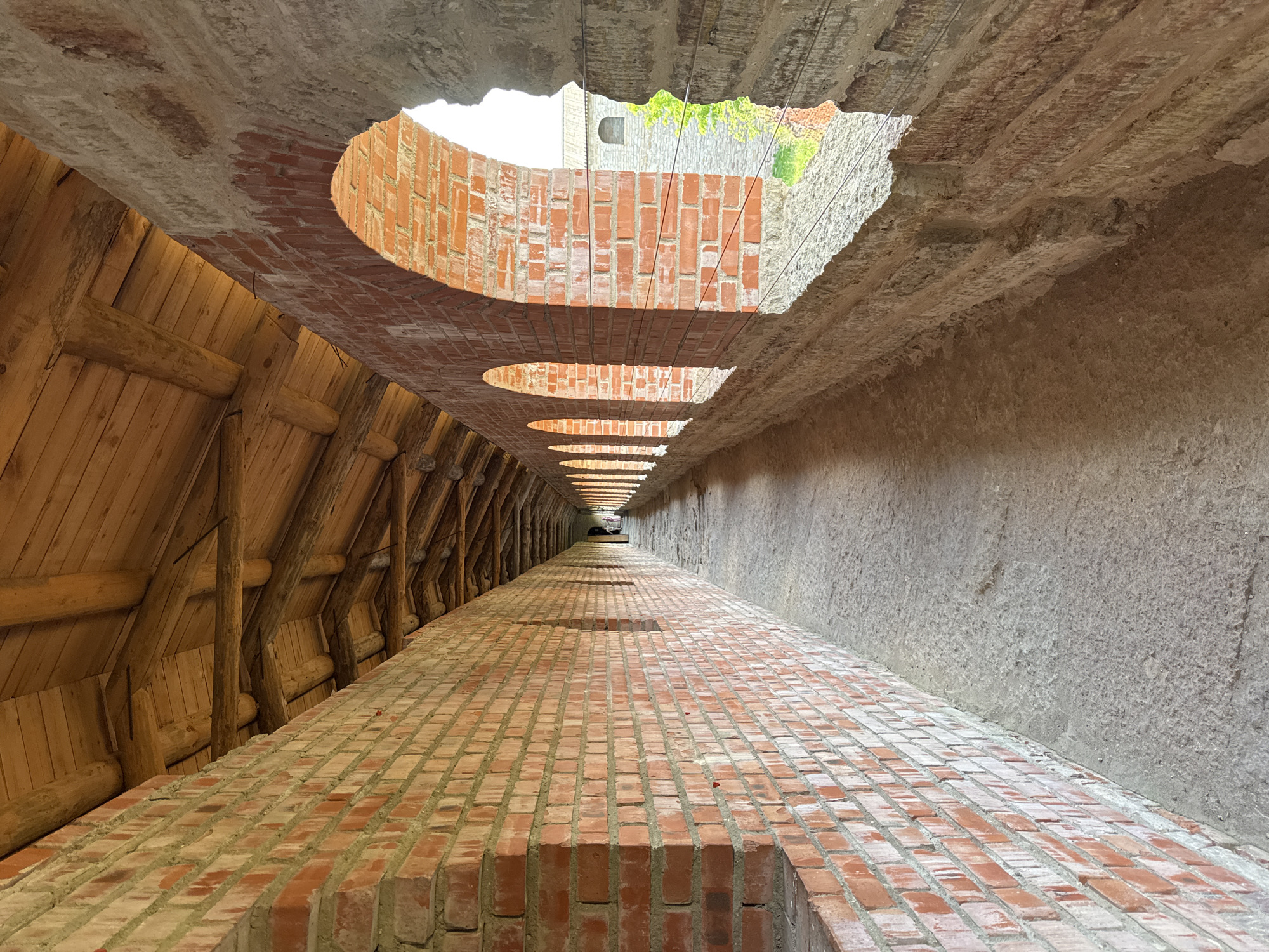 The image shows a long, narrow passageway with a brick floor and a rough concrete wall on the right. The ceiling is made of concrete with several oval-shaped openings that allow light to enter. On the left side of the passageway, there is a wooden structure composed of angled beams and planks. The perspective draws the viewer's eye towards a distant point where the passageway appears to open up. The scene is empty of people. The lighting suggests it is daytime, possibly sunny, given the bright light coming through the openings. The rough texture of the concrete and the brickwork indicate an outdoor or partially enclosed architectural structure, possibly an historical building or fortification given the context of Shlisselburg, Russia, a city known for its fortress. There is no visible text in the image.