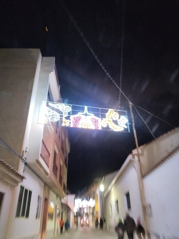A dimly lit street at night is framed by buildings on either side. Holiday decorations hang overhead, displaying illuminated designs in white, red, and yellow. Figures walk along the street, their forms blurred. Streetlights illuminate the path ahead, with more decorations visible further down. The sky is a dark backdrop, emphasizing the lighted elements in the foreground.