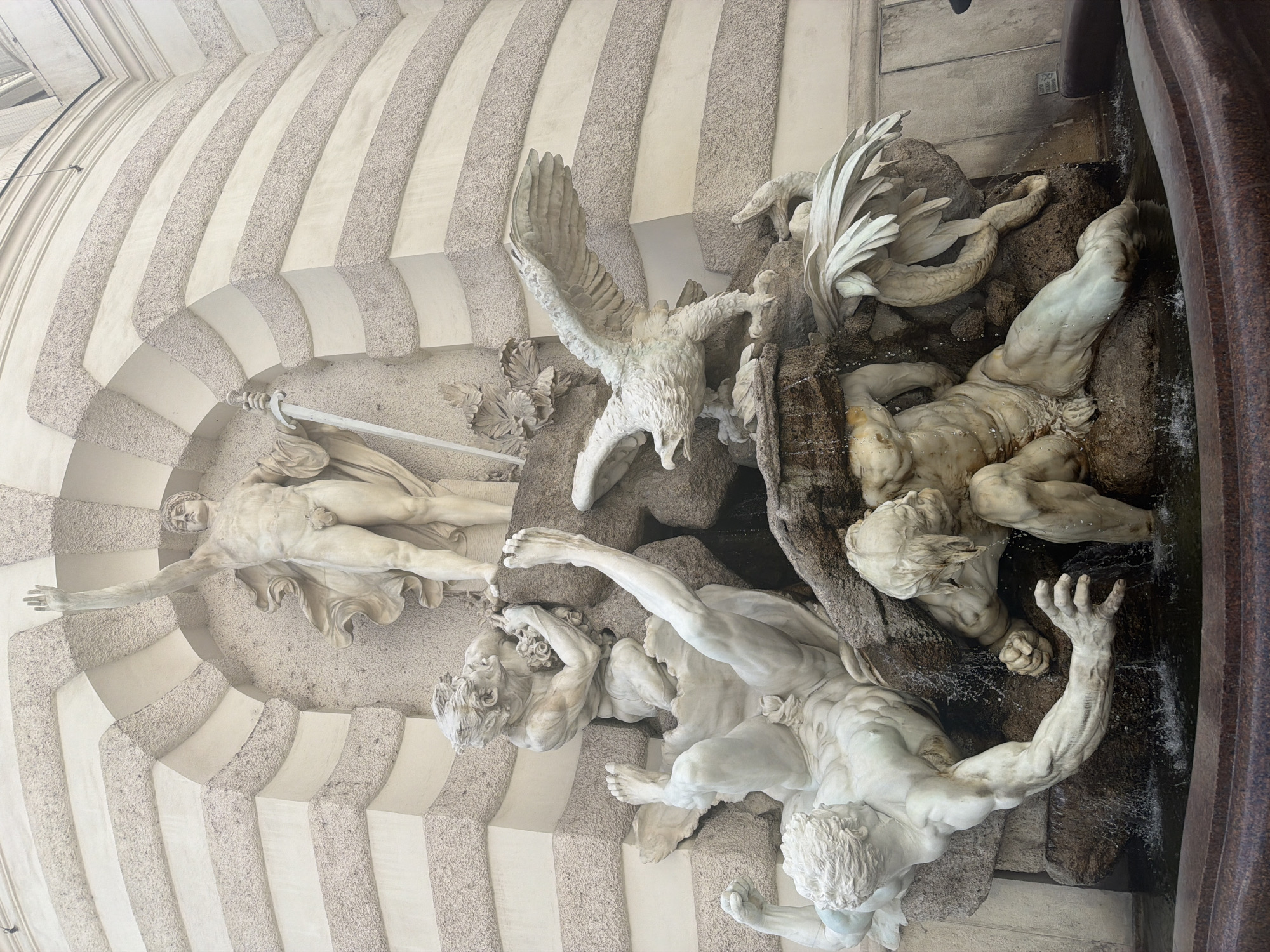 This media file captures a detailed view of the Pallas Athene Fountain in Michaelerplatz, Vienna, Austria. The scene features a complex arrangement of marble sculptures depicting mythological figures in dramatic poses. The central focus is Pallas Athene, a female figure standing tall and holding a spear, overseeing the action below.
Below Pallas Athene, several male figures are depicted in struggle and agony, entwined with serpents and rocks. An eagle sculpture is also prominent, with its wings spread, adding to the dynamic and turbulent atmosphere of the fountain. Water flows over the sculptures, creating a sense of movement and life within the frozen scene. The sculptures are intricately detailed, showcasing musculature and flowing drapery.
The setting appears to be an architectural space with a curved wall featuring alternating light and dark stone elements, suggesting it's part of a grand building or courtyard. The lighting suggests daytime, with no specific indication of weather conditions beyond the water flow. The overall mood is one of heroic struggle and divine power, common themes in classical mythology. No visible text is present in the image.
