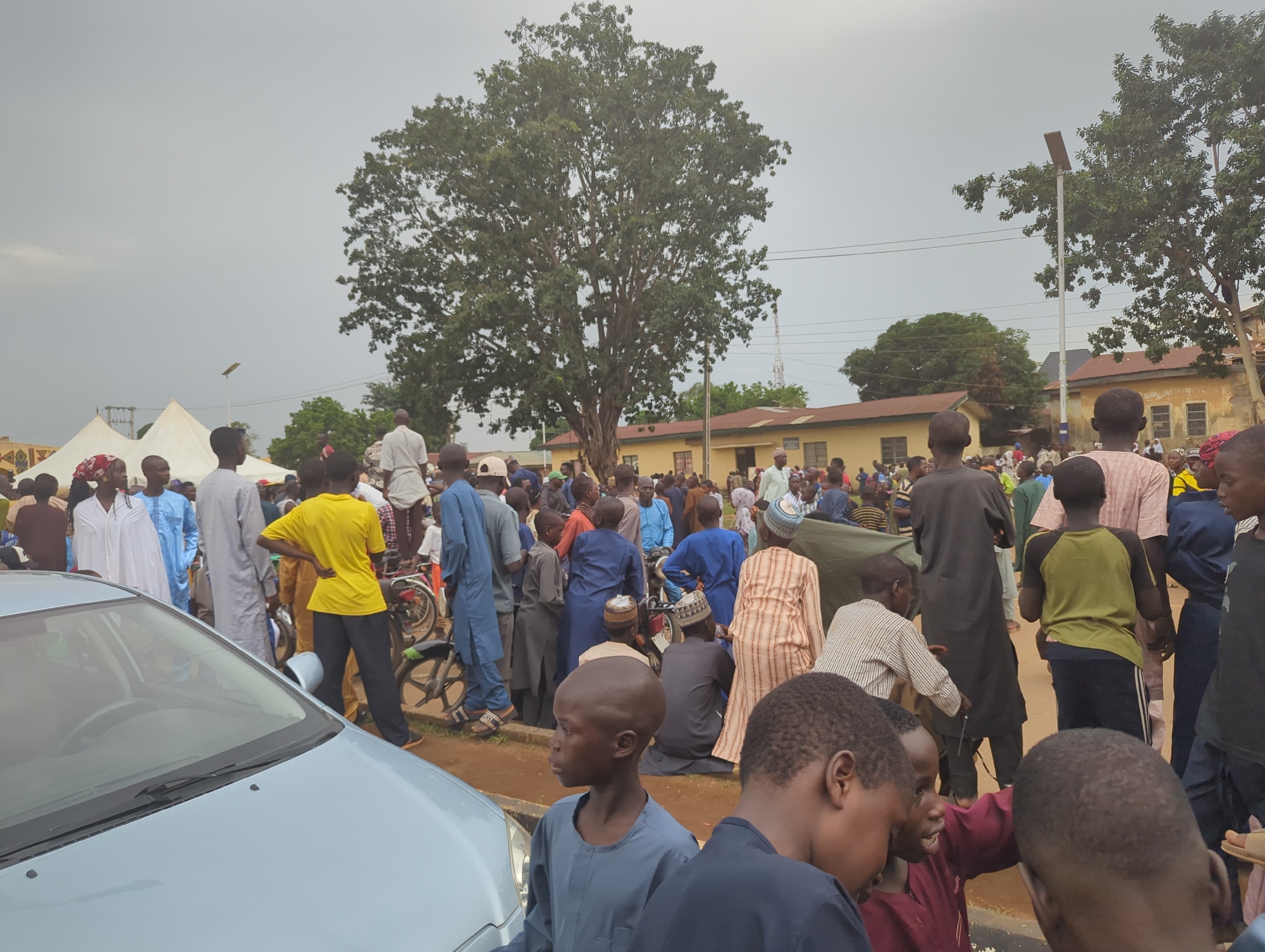 The image shows a daytime scene in Kafanchan A, Nigeria, with a gathering of people outdoors. The sky is overcast. A light blue car is prominently featured in the foreground on the left side of the image. A large tree is centered in the background. The crowd includes men, women, and children, many wearing traditional clothing such as long robes and head coverings. Some people are standing, while others are sitting. Bicycles are visible among the crowd. A single-story building with a brown roof is present in the background. There are utility poles and wires visible in the distance. Tents are set up in the background. The ground appears to be a combination of dirt and grass.