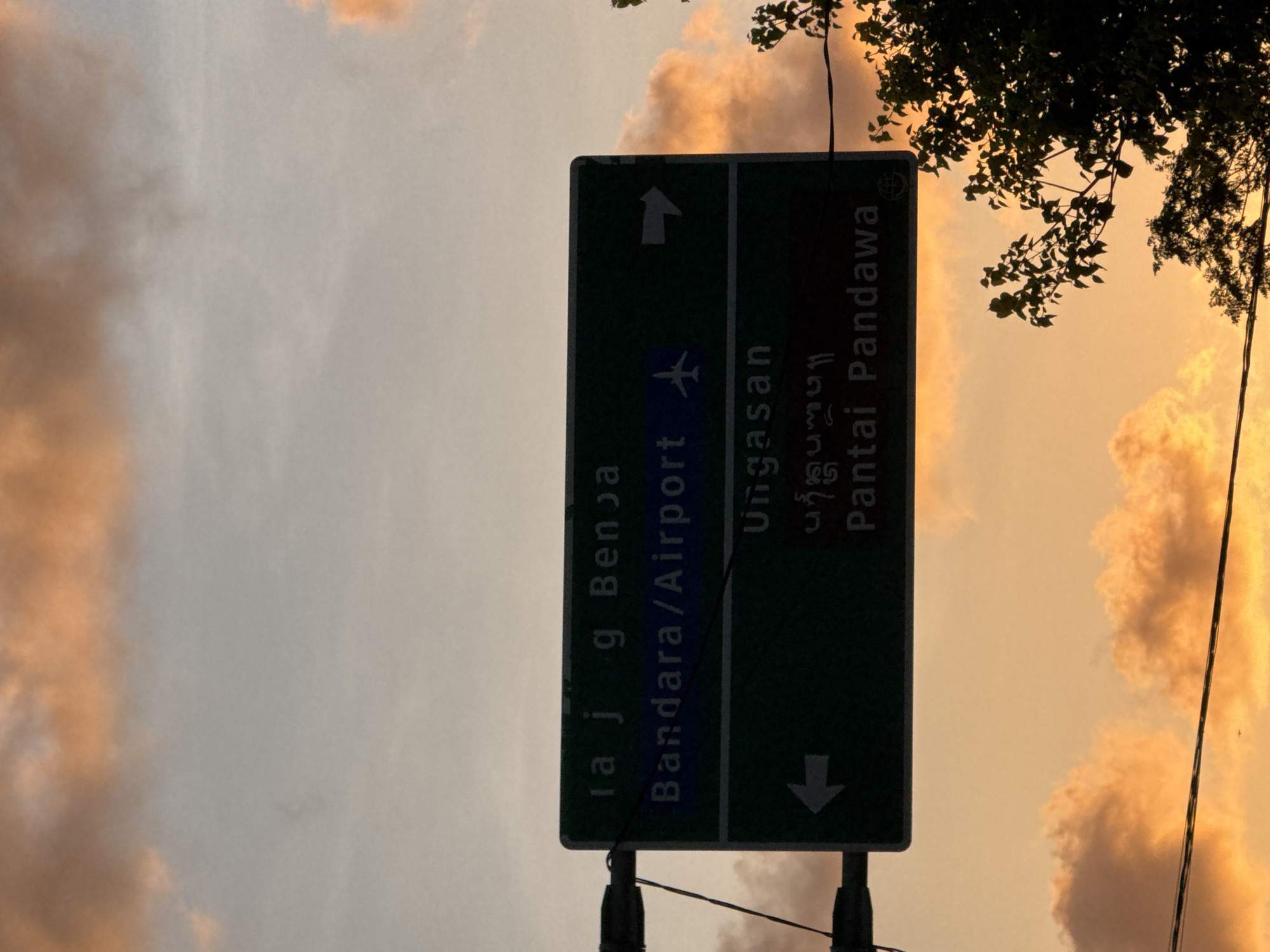 A green and white sign stands on a pole in Kuta Selatan, Indonesia, listing the directions to various places. The sign has an arrow pointing up next to the text "ra j g Benja" and an airplane icon next to the text "Bandara/Airport". The sign also includes an arrow pointing down next to the text "Ungasan" and the text "Pantai Pandawa". The background includes a pink and gray cloudy sky with the leaves of a tree in the upper right corner. Two wires also run through the bottom of the image in front of the sky.