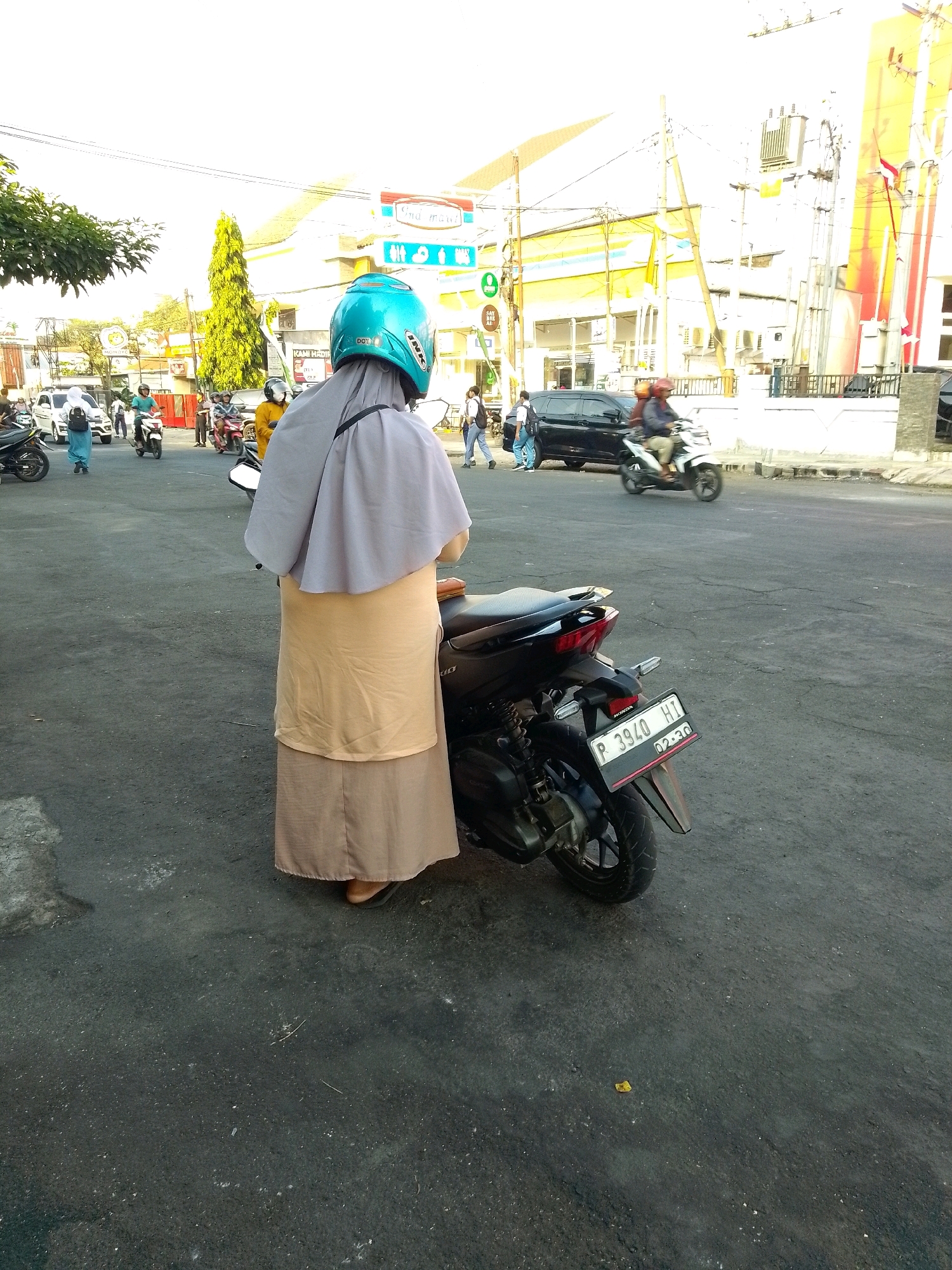 The image depicts a street scene in Jember, Indonesia, likely during daylight hours, given the bright sunlight. The focus is on a person from the back, wearing a light brown skirt and a grey hijab, with a turquoise helmet on their head. They are standing next to a black motorcycle, the rear of which is visible, including a license plate that reads "P 3940 HI".
The background reveals a busy urban environment. To the left, trees with green foliage are visible, with a tall, slender yellow tree in the mid-ground. Buildings with yellow and white facades dominate the background, suggesting commercial establishments. Various vehicles are present on the road, including a black car and several other motorcycles with riders. Some individuals are walking on the sidewalk. Electrical poles and wires are prominent throughout the scene. The road surface is asphalt, showing some wear and tear. The overall impression is one of a typical day in an Indonesian city, with traffic and pedestrian activity.