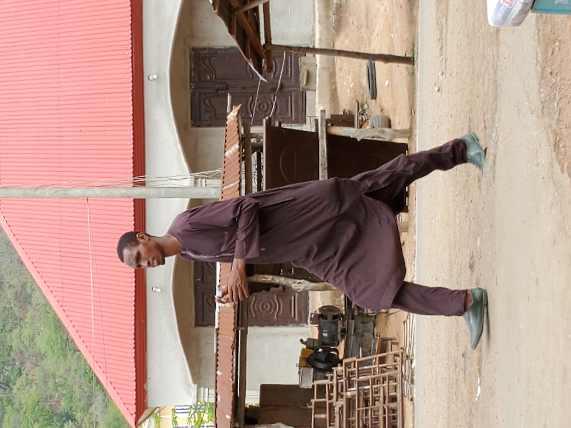 A man stands on a street in Mararaba, Nigeria, outside a building with a red roof. He wears a long, brown garment and green shoes. The building behind him has brown doors. In front of the building are metal frames and a yellow and black machine. The ground is dirt and gravel. A hill covered in trees is visible in the background.