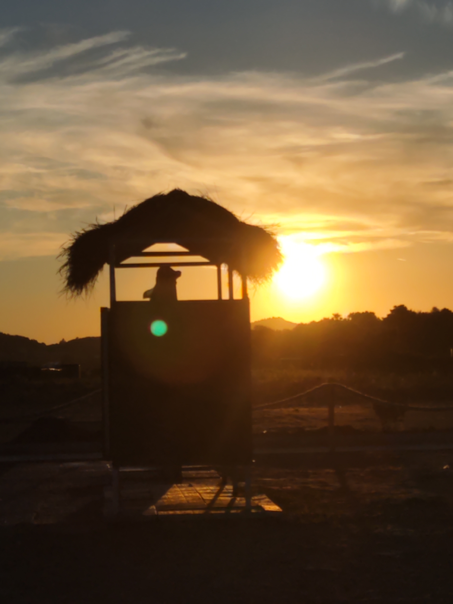 A silhouetted figure, possibly a person, stands within a thatched-roof structure at sunset. The structure appears to be a small, elevated booth or observation post. The sun is low on the horizon, casting a warm, orange glow across the sky and illuminating thin, wispy clouds. The foreground is dark and indistinct, with hints of ground and possibly a wooden platform. In the background, a silhouette of trees and hills is visible against the bright sky. The overall scene suggests a tranquil, natural setting. The specific location is Donji Štoj, Montenegro.