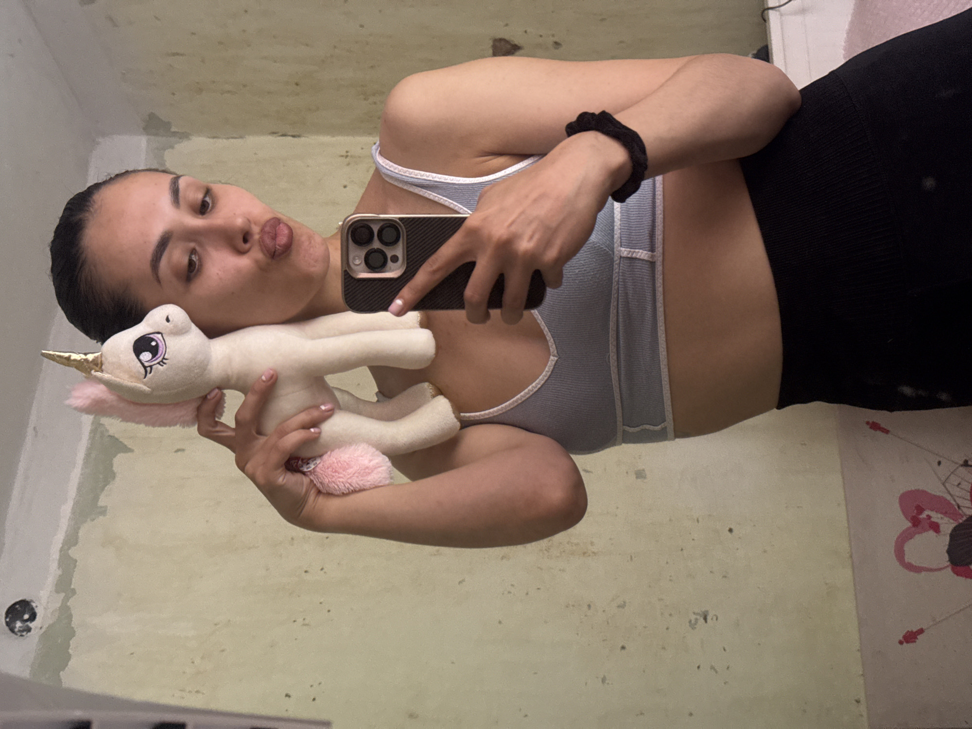 In Votkinsk, Russia, a young woman with dark hair pulled back is taking a selfie in a room with a textured, pale yellow wall. She is wearing a light blue sports bra and black leggings. She is holding a white plush unicorn with a pink mane and tail, and a gold horn, up to the side of her face, puckering her lips. The unicorn has large, stylized purple eyes with eyelashes. The woman is holding her smartphone, which has a dark case, in her right hand. A black scrunchie is visible on her left wrist. The wall behind her appears weathered and shows signs of repair or damage. On the lower part of the wall, a pink and red drawing of a heart is visible.