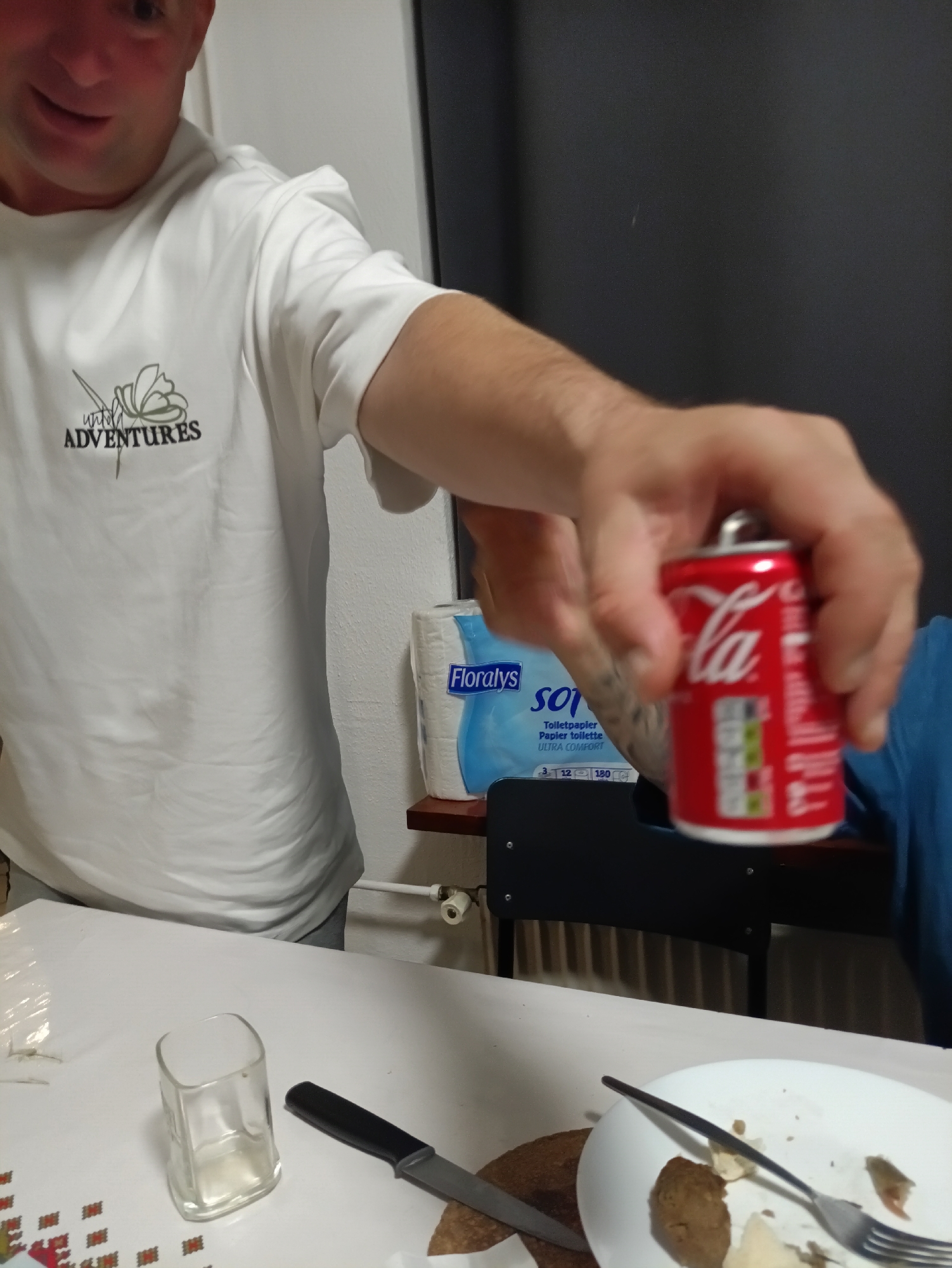 A person wearing a white t-shirt with a "ADVENTURES" logo is holding up a small can of Coca-Cola, offering it towards the camera. The person's face is partially visible, with a smile. In the background, a package of Floraly's toilet paper is visible. On a table in front of the person, there is an empty glass, a knife, and a dirty plate with food remnants and a fork. The setting appears to be indoors, possibly a dining area, with a dark wall in the background. The image does not provide any specific visual cues to identify the location as Zwartewaal, Netherlands.