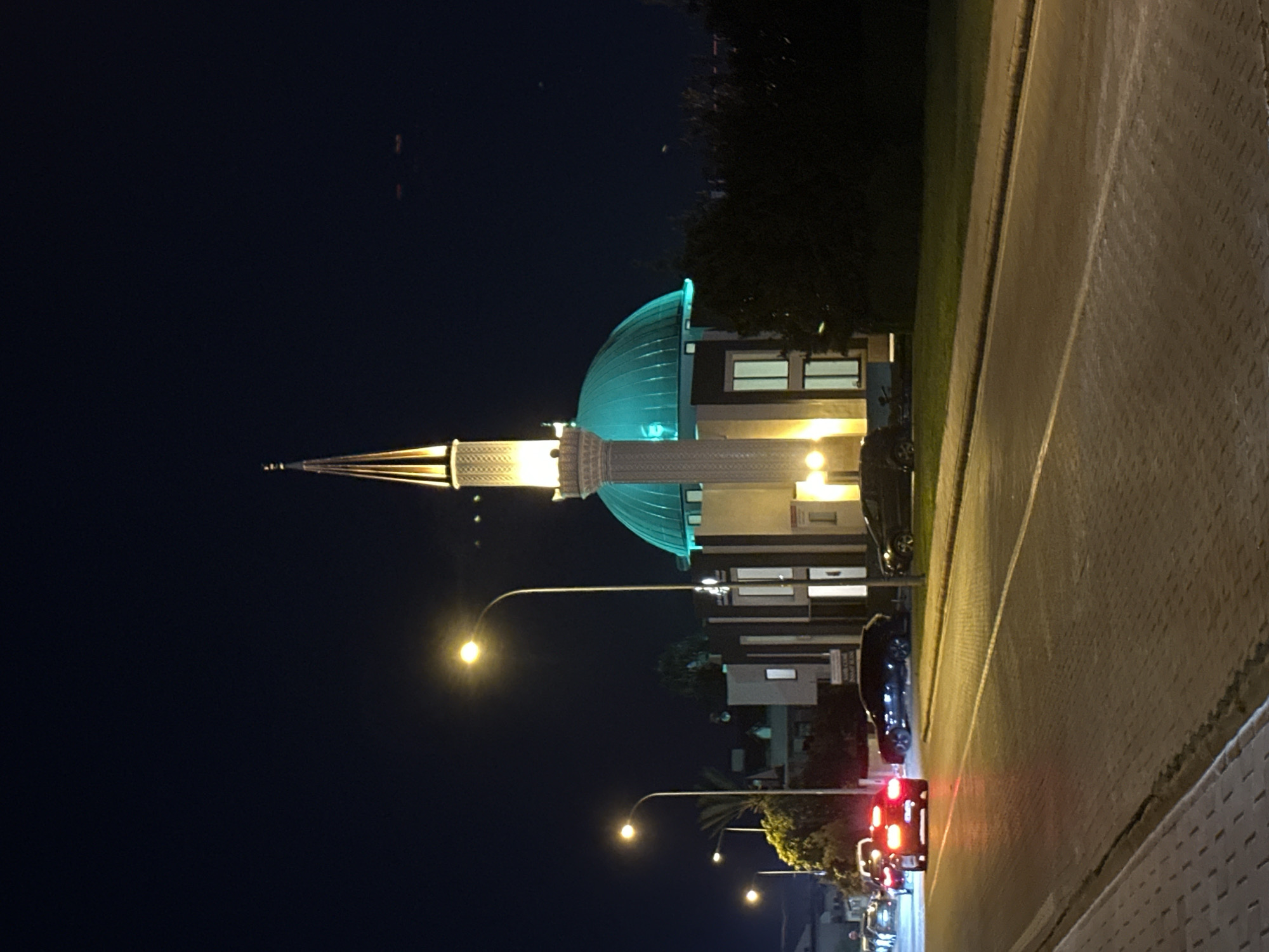 The image depicts a mosque at night in Fethiye, Turkey, likely the Fethiye Merkez Mosque. A minaret topped with a golden spire and a prominent turquoise dome dominate the structure. The mosque is illuminated, casting a warm glow on its facade. In the foreground, a paved road runs alongside the mosque, with several cars parked and driving. Streetlights line the road, illuminating the scene with their warm, yellow light. The sky above is dark, indicating it is nighttime. The overall scene is one of urban nighttime activity, with the mosque as the central architectural element.