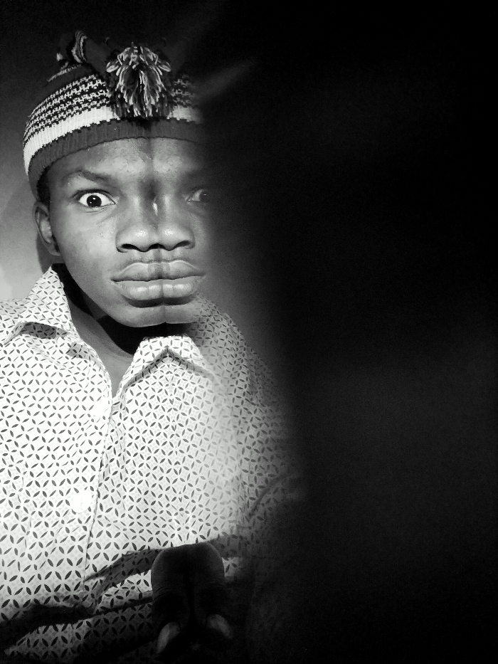 A black and white portrait captures a young man with a surprised expression, wearing a patterned shirt and a striped knit hat with a pom-pom. His face is partly obscured by a large, indistinct black shape dominating the right side of the frame. The stark contrast between light and shadow creates a dramatic effect, highlighting his wide eyes and pursed lips.
