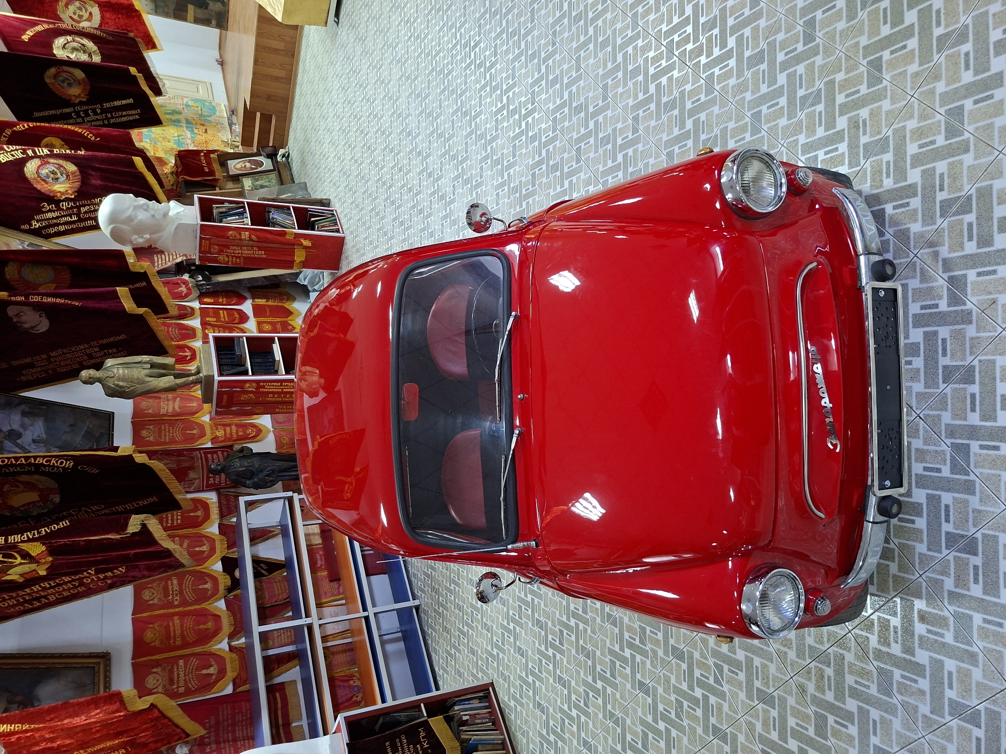 The image shows the interior of a museum in Ialoveni, Moldova, featuring a red vintage car as the central exhibit. The car is a small, compact vehicle, possibly from the Soviet era, with a rounded design and chrome accents. It is parked on a tiled floor with a geometric pattern.
In the background, there are displays related to Soviet history and ideology. Red banners with Cyrillic inscriptions and Soviet symbols hang from the walls. To the left of the car, there are white shelves filled with books and more banners. To the right of the car, there is a bust of a man, likely a political figure, placed on a red shelving unit. Further in the background, there are other exhibits including what appears to be a small stage and seating area. The overall impression is of a historical exhibition dedicated to the Soviet period. No people are visible in the image.