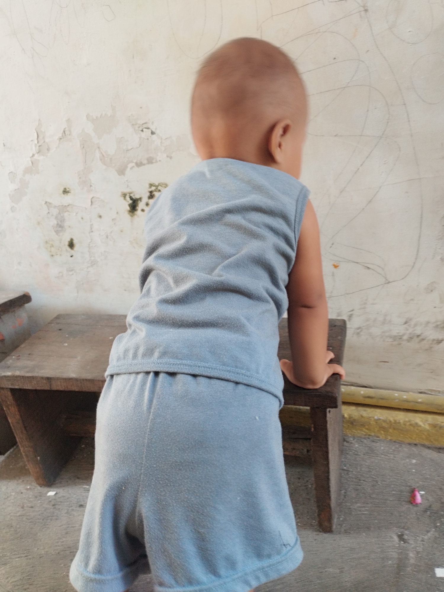A young child, seen from behind, stands on a wooden bench against a weathered wall in Surabaya, Indonesia. The child is wearing a light blue sleeveless shirt and matching shorts. The wall behind the child is dirty and cracked, with faint charcoal drawings and some green mold patches. The bench is made of dark wood and appears old and worn. The ground in front of the bench is concrete, with some debris visible.
