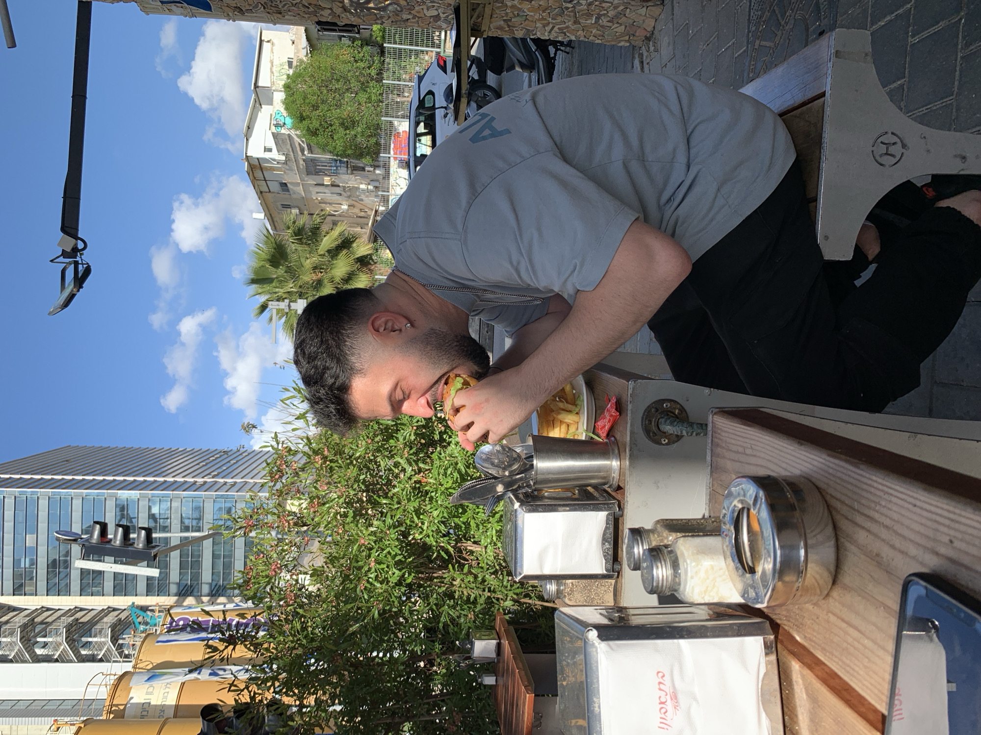A man with dark hair and a beard is bending over to eat a burger and fries at an outdoor table. The table is set with condiments and napkins. In the background, there is a green tree and a city street. The city is likely Tel Aviv, Israel, given the presence of a building with Hebrew writing in the background and the general architectural style.