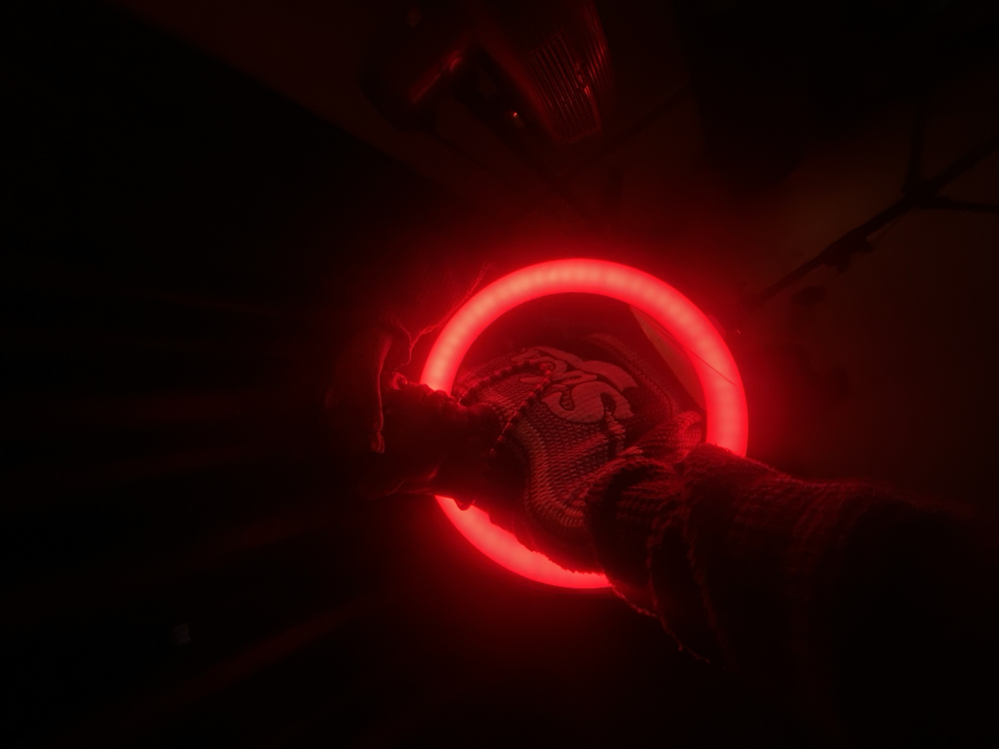 The image shows a close-up, low-angle shot of a person's arm and hand holding a glowing red ring light against a dark background. The person is wearing a textured sweater or jacket and a necklace. The ring light is the primary light source, casting a strong red glow on the arm and hand. The background is mostly black, with some subtle red light rays emanating from the ring. To the right, a dark object with horizontal lines, possibly a speaker or vent, is faintly visible. The overall mood is dramatic and mysterious due to the low lighting and the intense red glow. The scene appears to be indoors, in a dimly lit studio or room. There is no visible indication of the specific location in Abakaliki, Nigeria.