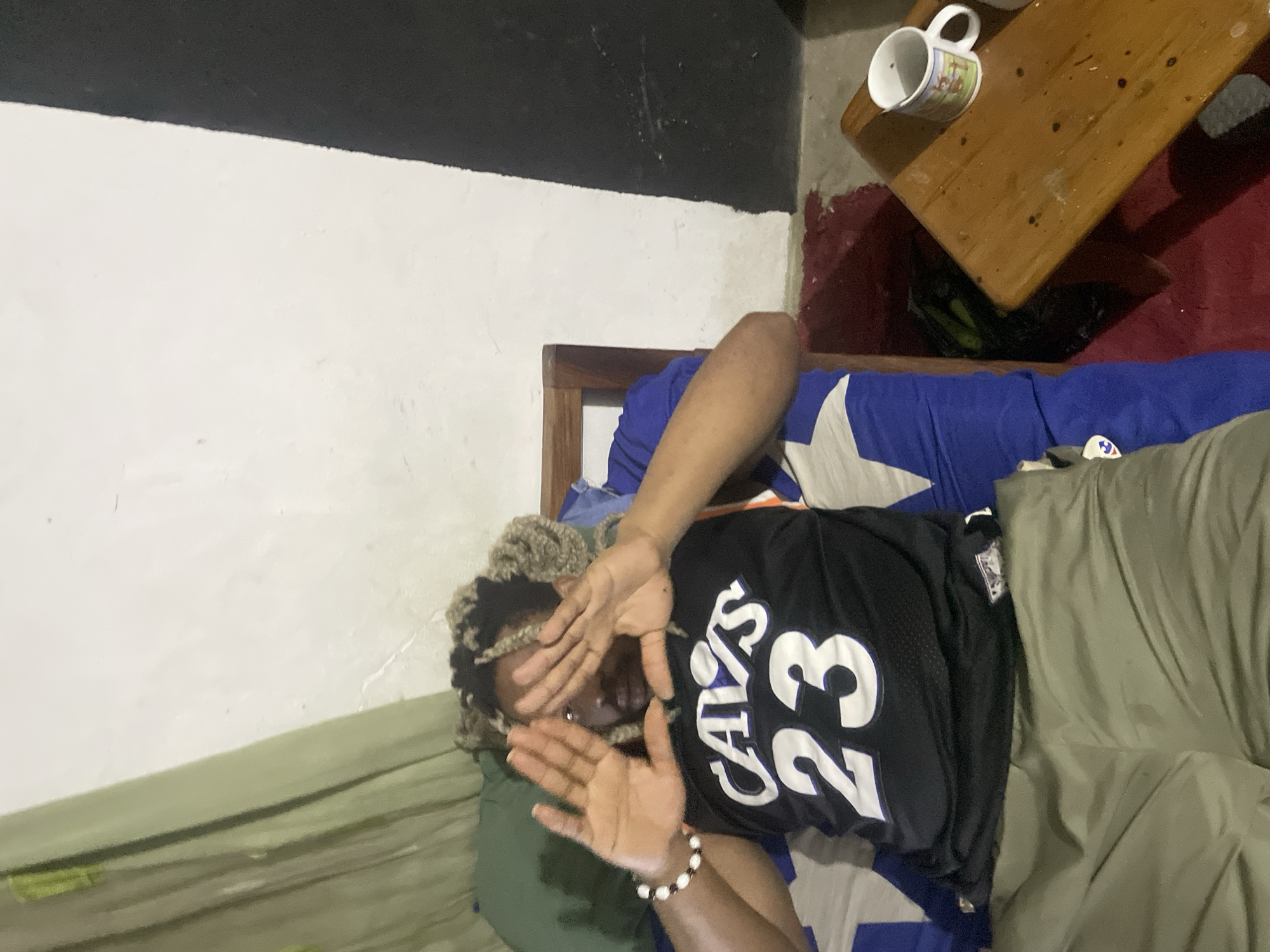 A person with blonde hair is lying on a bed. They are wearing a black "Cavs 23" jersey, light green pants, and a bracelet. Their hands are raised, partially covering their face. The bed has a blue and white patterned sheet. The room has white walls with a black band near the ceiling. A wooden bedside table with a mug on it is visible in the background. The scene is indoors. The location is Jos, Nigeria.