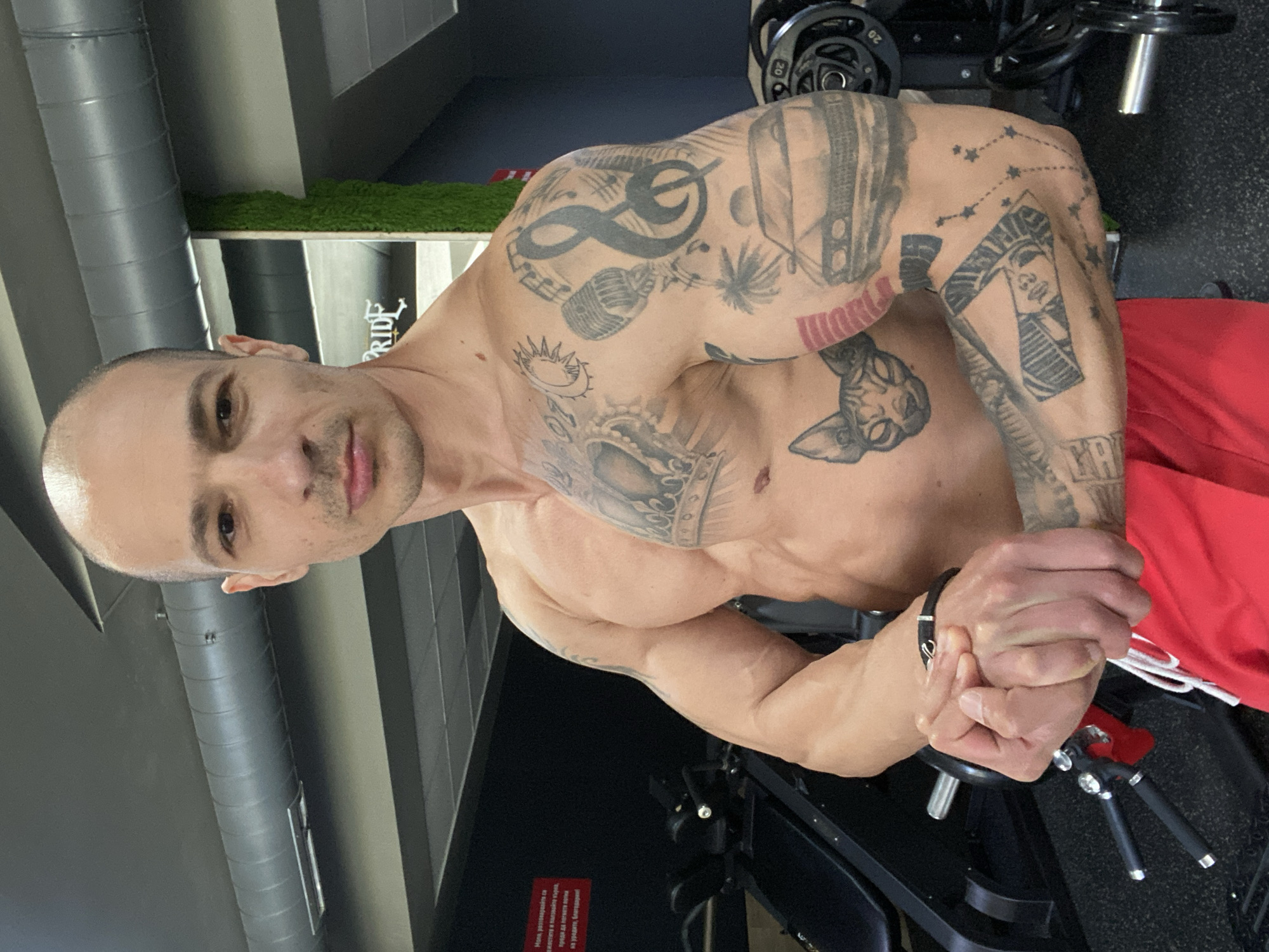 A shirtless man with a shaved head is standing in what appears to be a gym or workout area. He has numerous tattoos on both arms and his torso. He is wearing red shorts with white drawstrings and a black wristband on his left wrist. His hands are clasped together in front of him. Behind him are weight plates, part of a weight machine, and flooring that looks like it could be in a gym. In the background is a sign that says "PRIDE" and what appears to be some decorative artificial grass. The city is Pleven, Bulgaria, based on the visible text in a gym's signage.