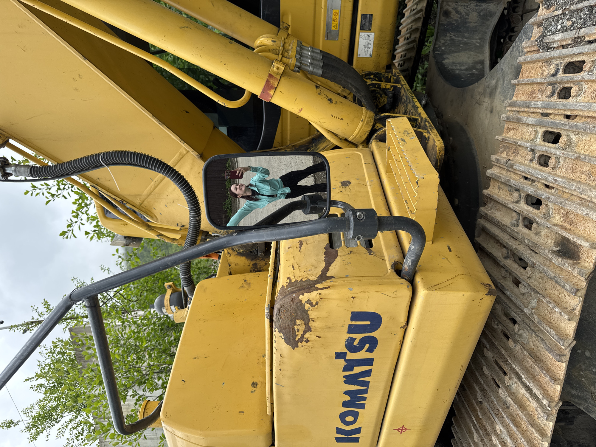 The image shows a yellow Komatsu excavator with a woman reflected in its side mirror. The excavator is likely located in Krasnaya Polyana, Russia. The woman is holding a mobile phone and taking a selfie in the mirror. The excavator is positioned on a surface that might be gravel or asphalt. The background includes trees and a building under a cloudy sky. The excavator's tracks are heavily worn and possibly rusted.