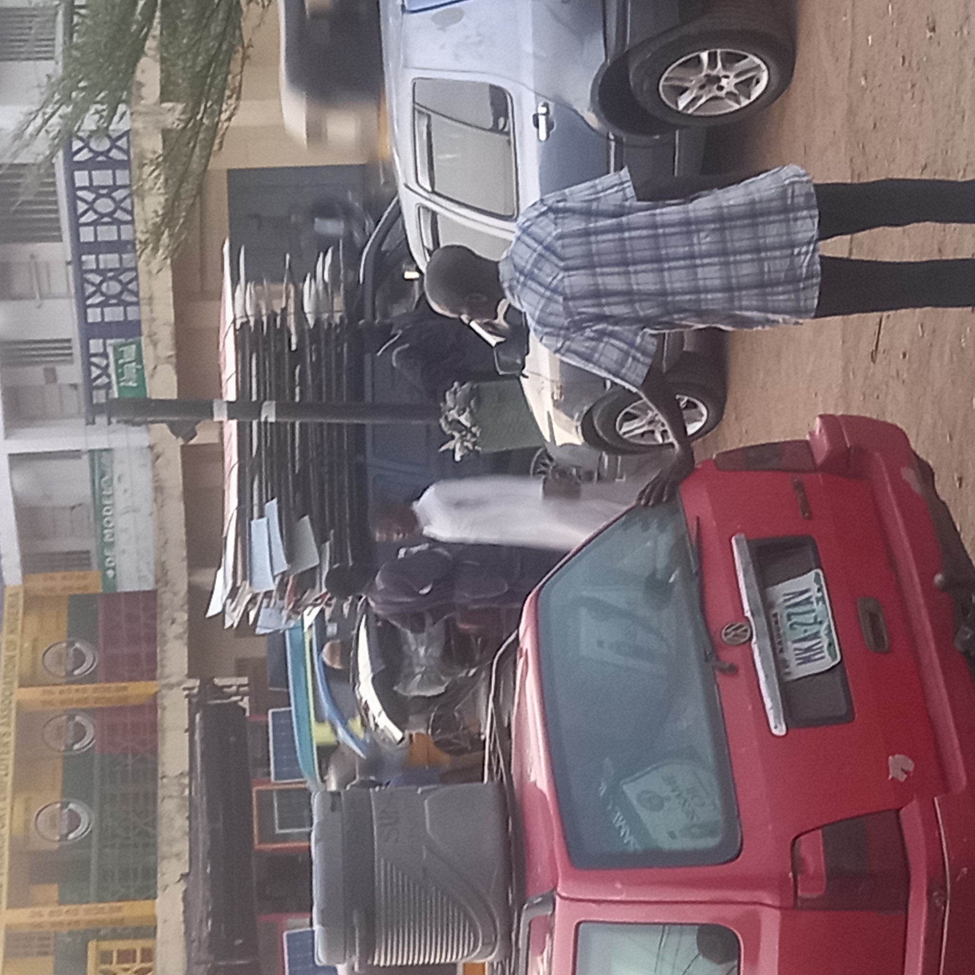 The image shows a street scene in Dadi Riba, Nigeria. A red car is in the foreground with license plate "NKA-27AV". A man is standing next to a car wearing a blue and white checkered shirt and black pants. A white cloth is visible behind the red car. Additional cars are in the background. A building is visible with signage. Crates stacked next to a wall can be seen on the building's facade. Multiple people are present near a van loaded with materials.