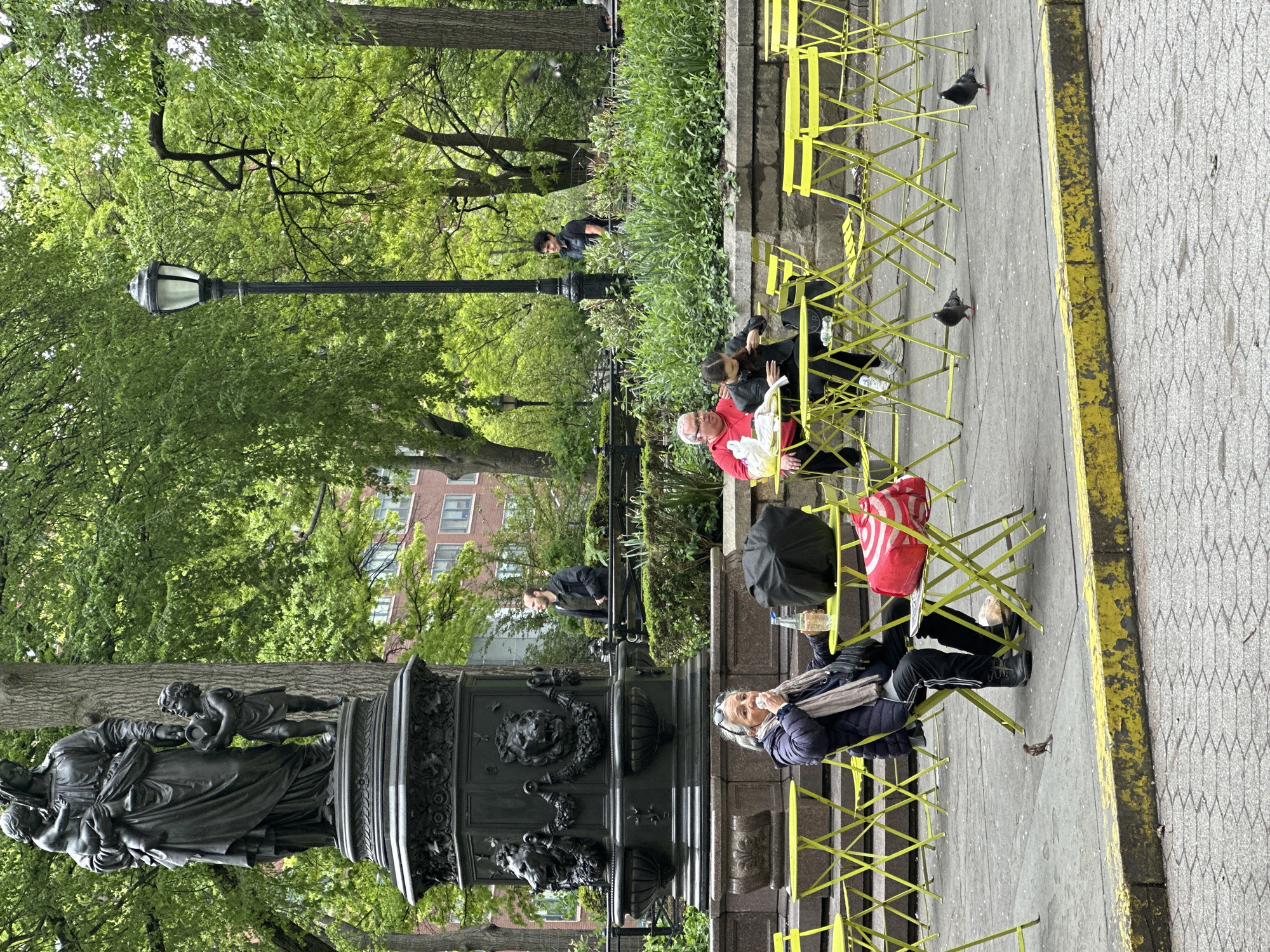 This outdoor image depicts a lively urban park scene in New York, United States of America, under bright, possibly overcast, daytime conditions. The setting features a paved walkway bordered by a low wall and a line of bright yellow folding chairs. Several people are seated on these chairs, appearing relaxed. An older man in a red shirt is seated alongside a woman in a black top, both engaged in conversation. Further down the row, an older woman with grey hair and glasses, wearing a blue jacket and scarf, looks up while holding a plastic bottle and wiping her mouth. A red and white Target shopping bag is placed on one of the chairs, and a black umbrella rests on another.
To the left, a prominent dark bronze monument stands, featuring classical figures including a woman holding two children and another standing child. Its base is intricately decorated with lion heads and floral motifs. A tall, classic black lamppost is positioned in the mid-ground. The park is characterized by lush green trees with abundant foliage, indicating spring or summer. Through the dense greenery, brick buildings are visible in the background, affirming the urban setting. Pigeons and a small bird are seen on the paved ground near the chairs, with some appearing to forage. Other individuals are scattered throughout the park, some near the monument and others among the trees. The overall atmosphere suggests a casual day in the park.