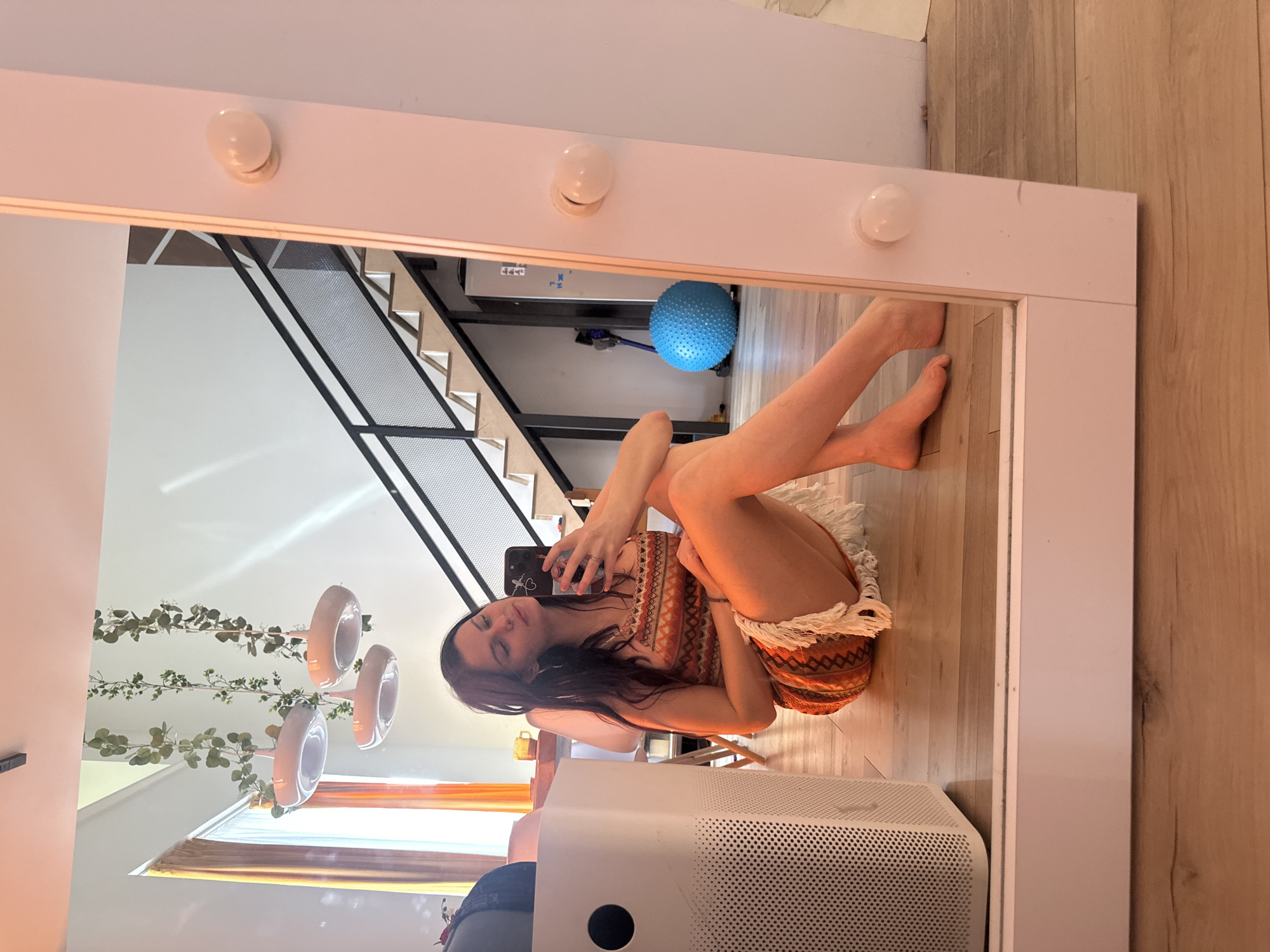A woman sits on the floor in front of a large mirror, taking a selfie. She has dark hair and is wearing a colorful, fringed dress. In the mirror's reflection, a staircase with a black railing is visible, along with a blue exercise ball and a white air purifier. The room appears to be a modern living space with wooden floors and high ceilings. The lighting suggests it is daytime.