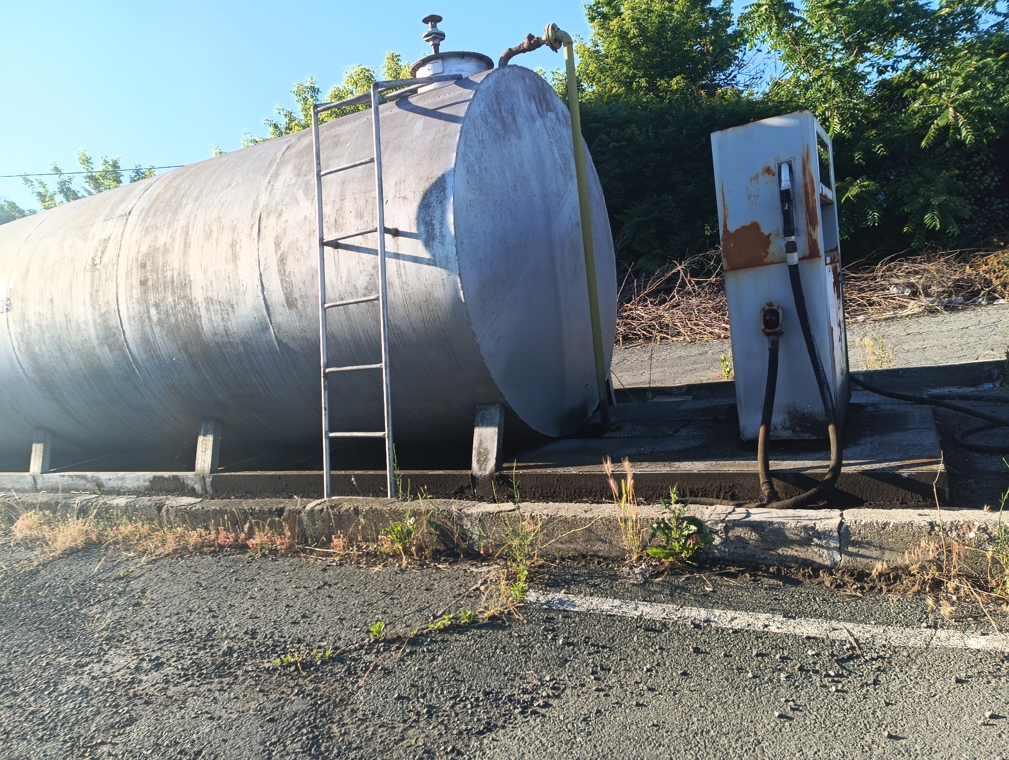 An outdoor scene in the city of Karnobat, Bulgaria, features a large, silver, cylindrical storage tank with a ladder on its side, positioned next to a rusty, weathered fuel dispenser. The tank appears to be made of metal and shows signs of age and exposure to the elements, with streaks of dirt and discoloration visible on its surface. The fuel dispenser, also metallic, has a dark hose connected to it and a nozzle hanging down. The ground around the tank and dispenser is asphalt, with scattered debris and dry vegetation growing along the curb. In the background, a dense line of green trees forms a backdrop against a clear blue sky, suggesting it is a sunny day. The overall impression is one of an industrial or storage area that is perhaps no longer in active use, given the state of disrepair of the fuel dispenser.