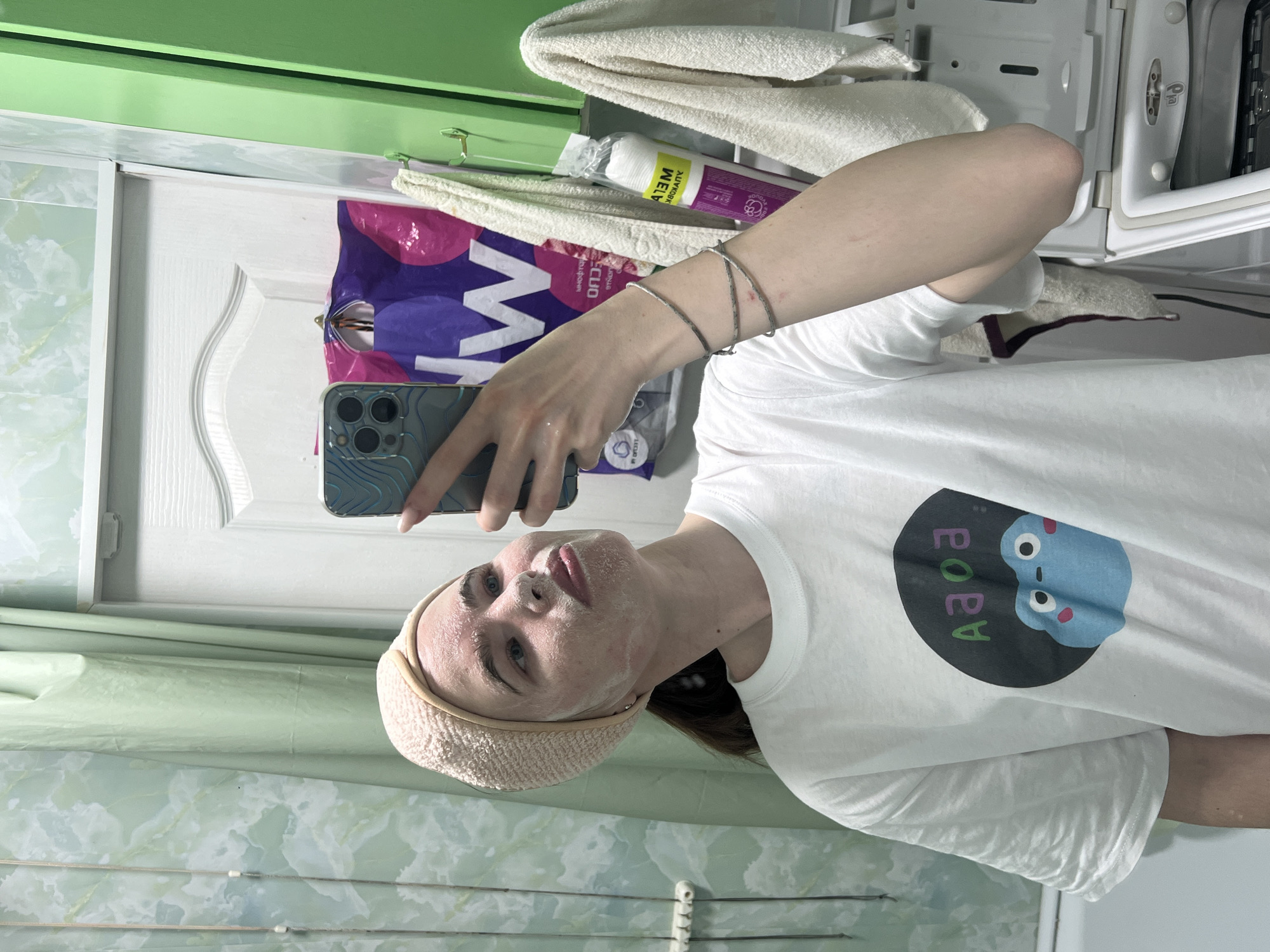 A person is taking a selfie in a bathroom in Chaikovsky, Russia. The person is wearing a white t-shirt with a cartoon graphic, a light-colored headband, and has a facial mask applied. They are holding a smartphone in their left hand. The background includes a white door with a decorative panel, green and white patterned bathroom tiles, a stack of white towels, various toiletries including one in a purple package, and a white appliance that appears to be a washing machine or dryer. The overall lighting is bright.
