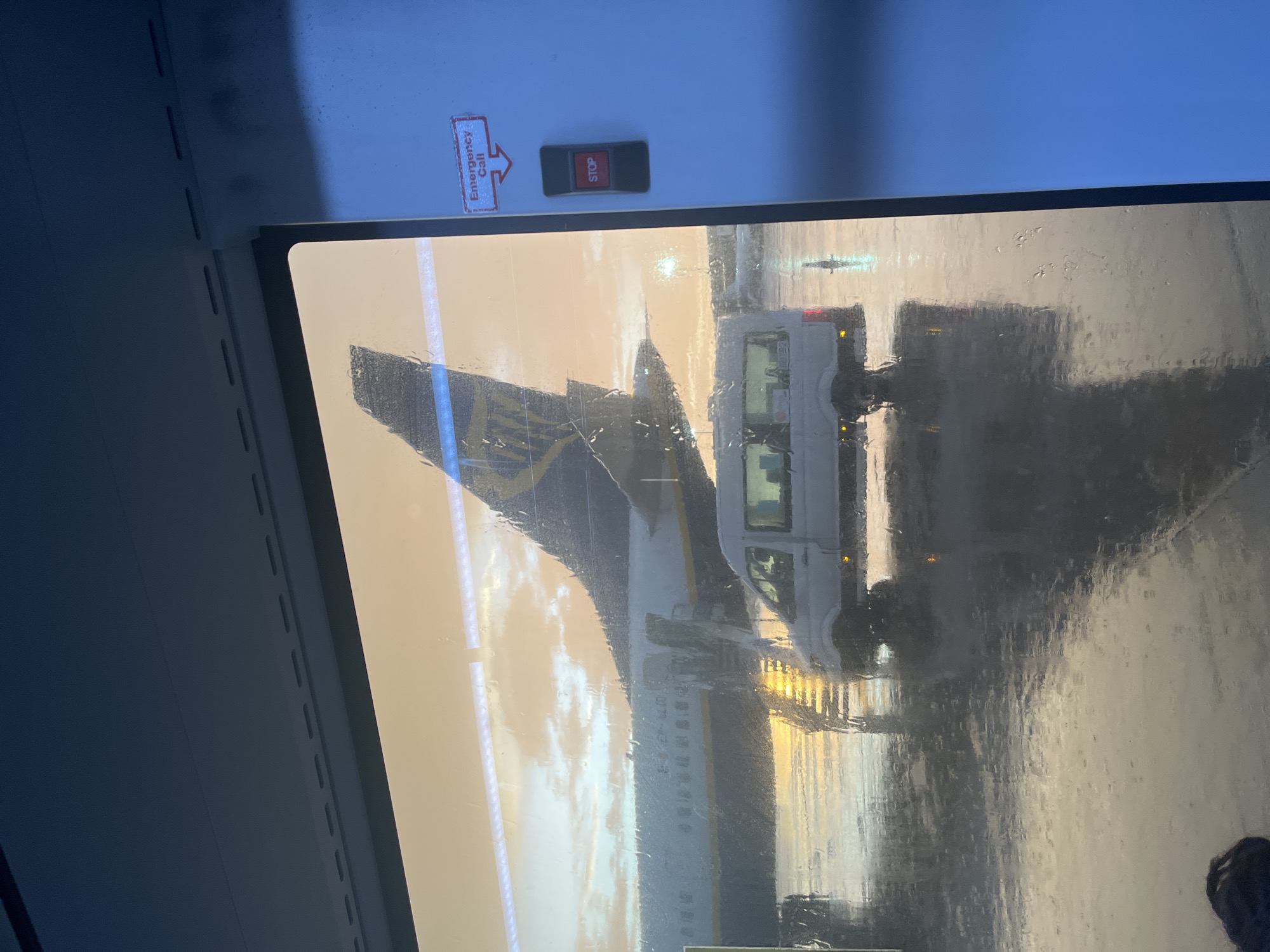 The image is taken from inside a vehicle, looking through a rain-streaked window. Outside, an airplane is visible, specifically its tail fin, which features the Ryanair livery. A white service van is also present. The presence of an airplane suggests the location is an airport. The text on the emergency call button is difficult to read, but the presence of Ryanair points to the possibility of Alicante–Elche Miguel Hernández Airport, located in the city of Elx / Elche, Spain. The light suggests either sunrise or sunset. There are no visible people in the image aside from a possible head at the very bottom-right.