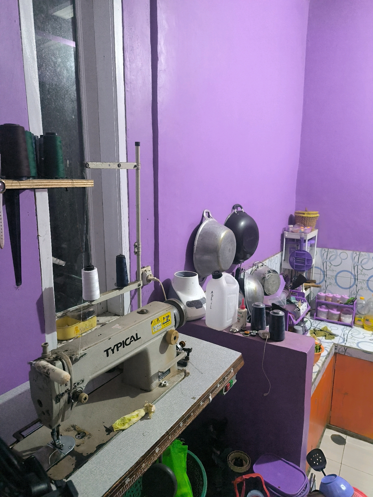 The image depicts an interior scene, likely a workspace or kitchen area, painted in a vibrant purple. In the foreground, a well-used industrial sewing machine, labeled "TYPICAL", sits on a table. Various spools of thread, some dark brown and green, are visible on a shelf above and to the left of the sewing machine, along with a white spool of thread on a stand next to the machine. To the right of the sewing machine, a counter area is visible, cluttered with kitchenware including several pots and pans hanging on the wall, a large white plastic jug, and a multi-tiered shelf holding small containers, possibly spices. A portion of an orange cabinet is visible in the far right. The overall impression is of a functional, possibly domestic or small business environment in Cilengkrang, Indonesia.