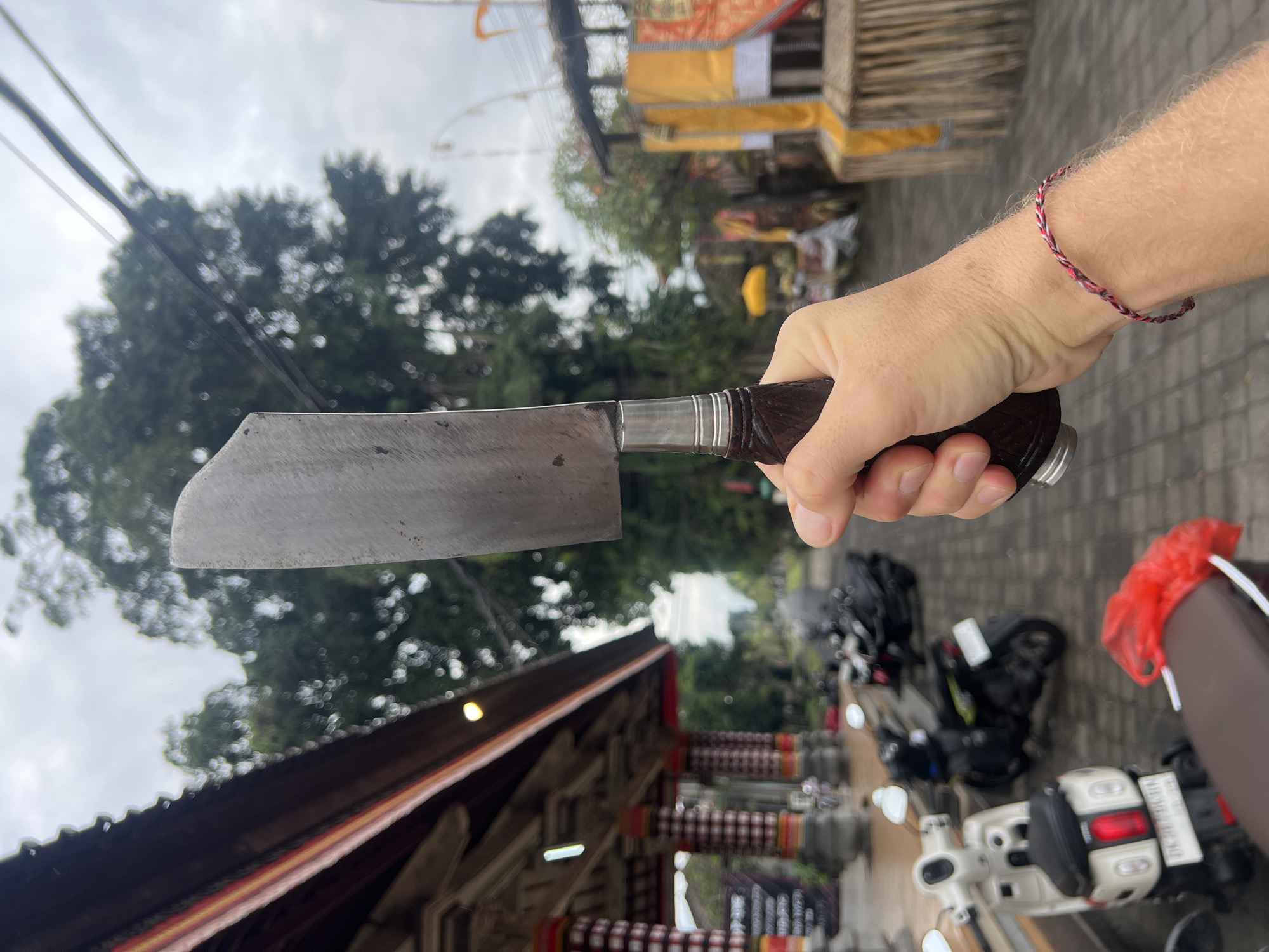 A right hand holding a large, dull cleaver with a brown handle is prominently displayed. A red and white braided bracelet adorns the wrist of the hand. The background features a Balinese building with a tiled roof and ornate decorations. A street scene includes parked motorbikes. The location is Sukawati, Indonesia. The cleaver is held in a potentially threatening manner.
