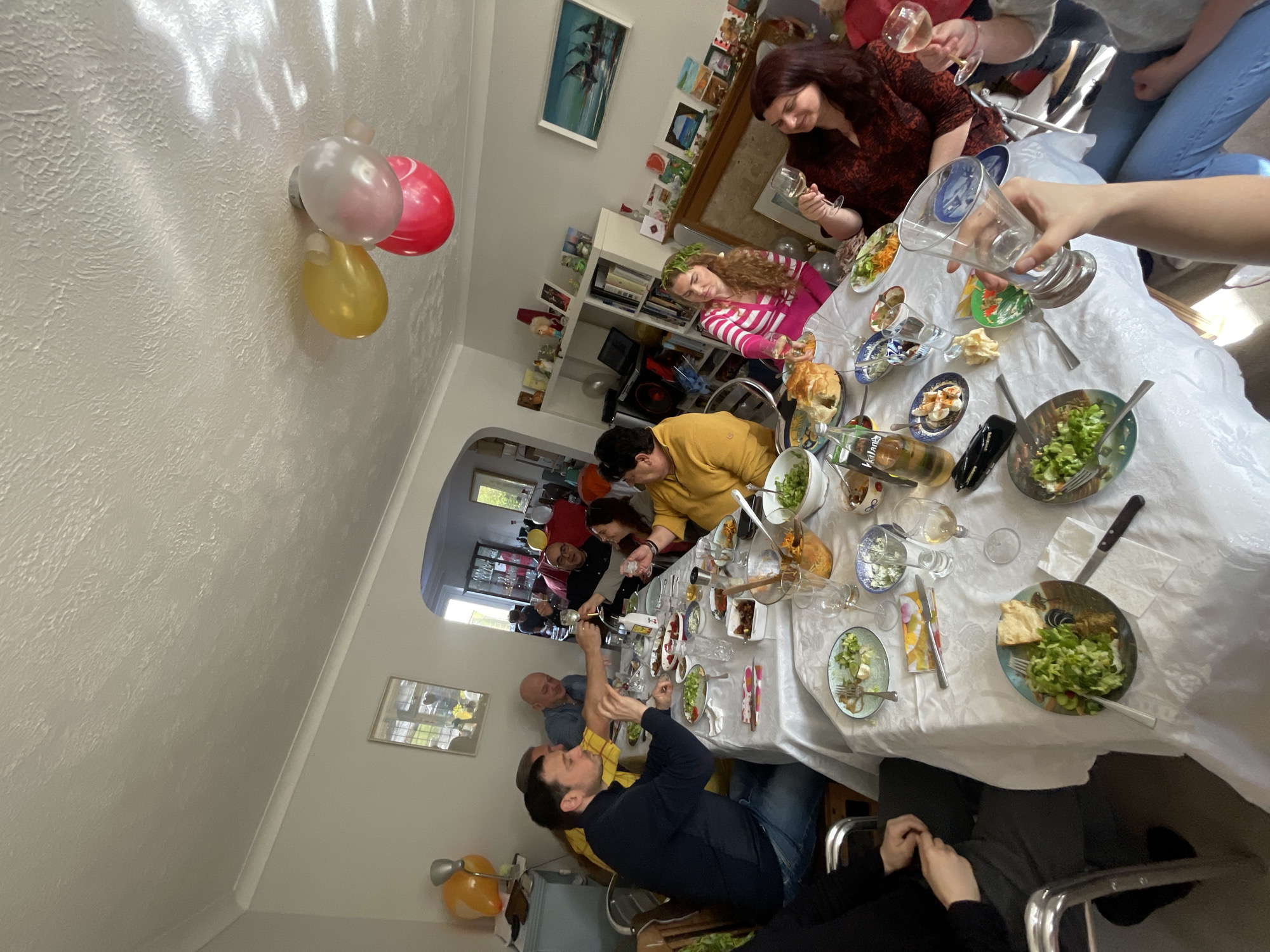 A gathering of people is seated around a table laden with food and drinks, seemingly in the midst of a celebratory meal. The table is covered with a white tablecloth and scattered with plates, bowls, cutlery, and glassware. Several individuals are holding glasses, some appearing to be engaged in a toast. The people present vary in age and are dressed in casual clothing. The background shows a home interior, with bookshelves and framed pictures on the walls. Balloons are attached to the ceiling. The location appears to be Maidstone, United Kingdom, based on visual elements and architectural features typical of residential areas.