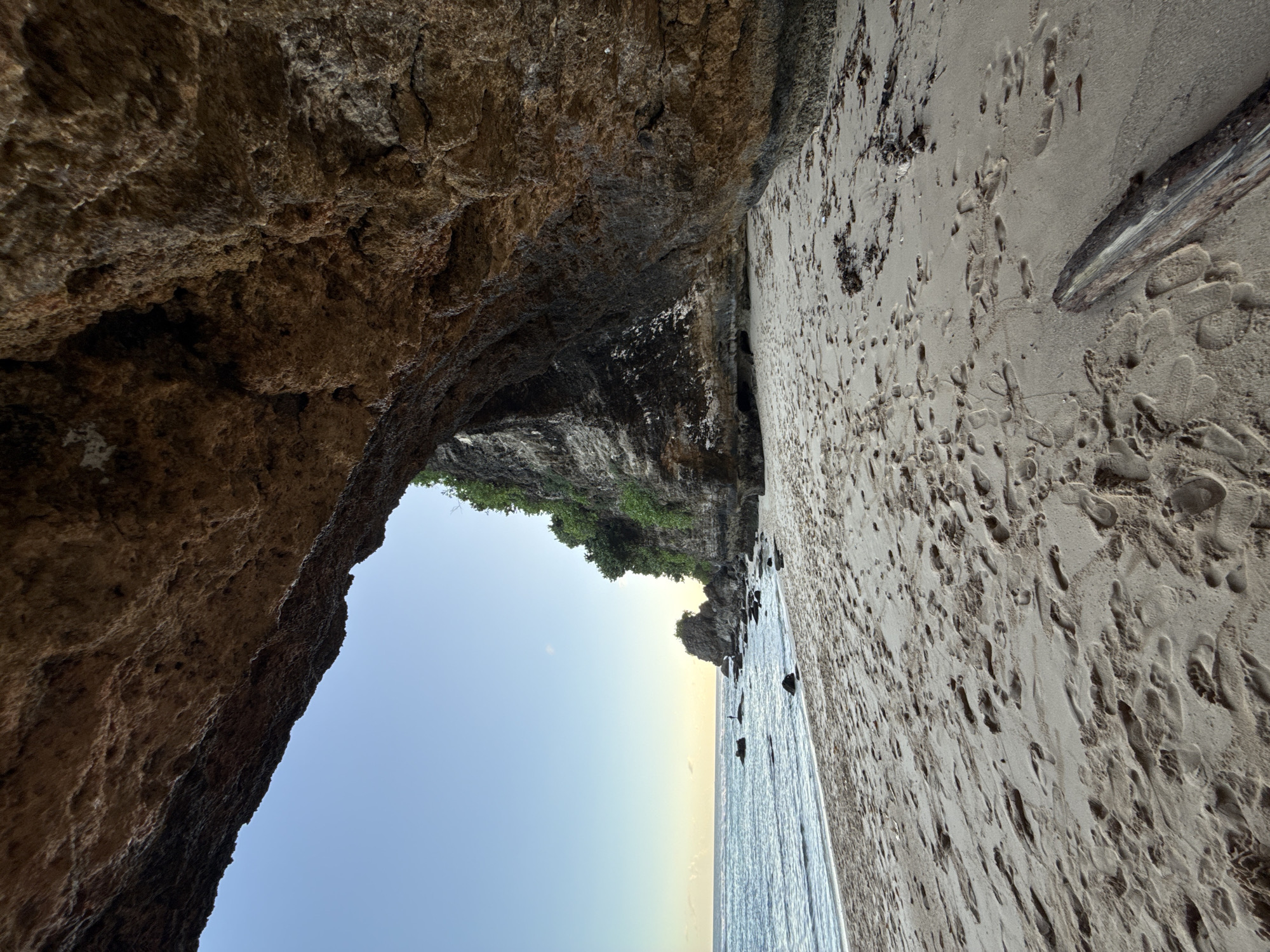 This photograph, taken in Ungasan, Indonesia, showcases a beach scene framed by a natural rock archway. The foreground features coarse-grained sand densely marked with numerous footprints, indicating heavy foot traffic. A large, weathered piece of driftwood lies on the beach surface. The backdrop is the ocean meeting the horizon under a clear sky. The archway consists of dark, textured rock formations covered with vegetation on its upper edges. The rock texture suggests sandstone. The water edge along the ocean is lined with small rocks.