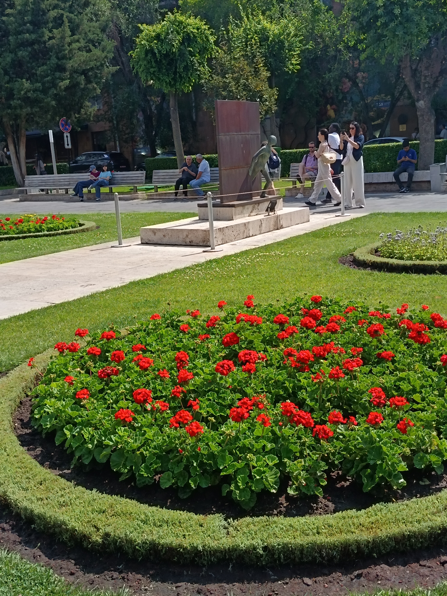 In this bright, sunlit outdoor scene, a manicured park area features a prominent circular flower bed filled with vibrant red geraniums and bordered by neatly trimmed green hedges. Beyond this, a concrete pathway winds through a lush green lawn. Further in the background, a group of people are seen enjoying the park. Some are seated on park benches, engaged in conversation, while others stroll along the path. A bronze statue of a boy is visible near a large, reddish-brown structure, possibly part of a monument or art installation. The area is surrounded by mature trees, casting shadows and adding to the serene atmosphere. A blue sign with a red circle and a white arrow is also present on a post.