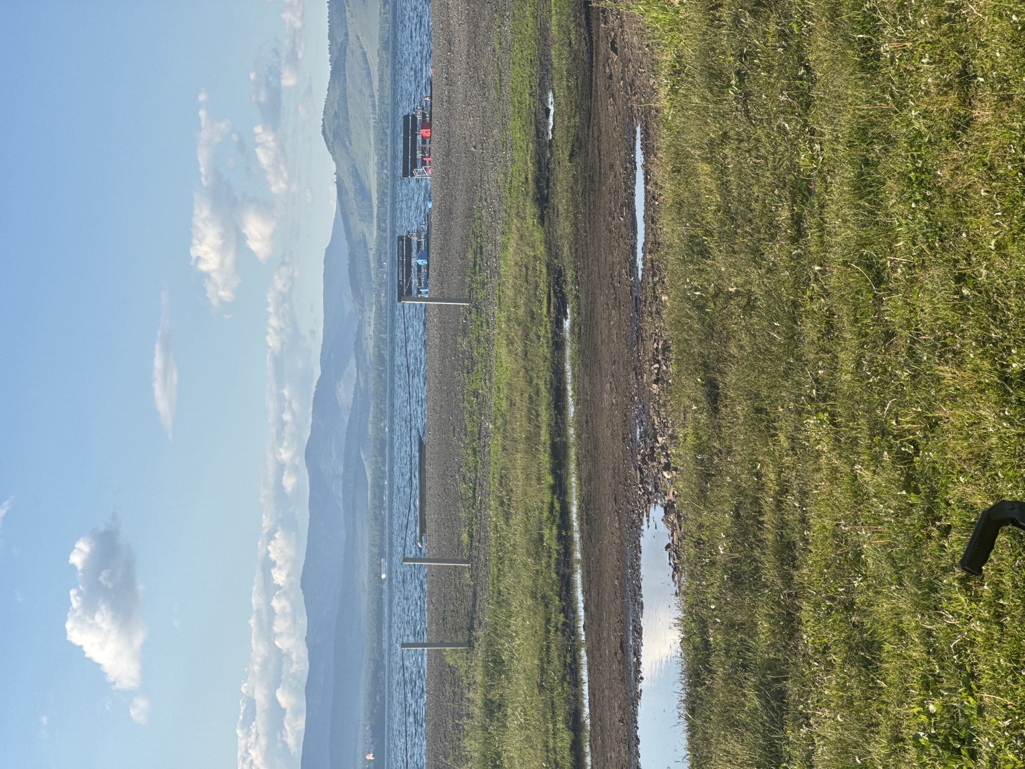 This is an outdoor, eye-level, full shot image of a lake and its surroundings. The sky is a light blue with scattered white clouds, some of which are large and puffy. In the middle ground, a body of water with choppy waves extends across the frame, meeting a pebbly shore. Two docks or platforms with benches are visible on the water. The foreground is dominated by green grass, with a muddy path and puddles of water closer to the shore. In the far distance, rolling hills with a faint green hue are visible under the hazy sky. The scene appears to be located near the city of Якты-Куль, Russia, possibly a recreational area by a lake.