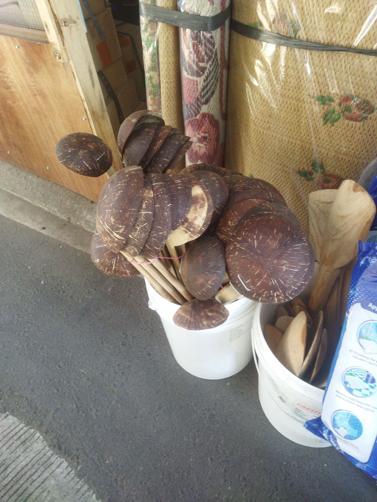 In a shallow depth of field, a collection of coconut shell bowls and wooden utensils are displayed on a gray concrete surface. The coconut shell bowls, stacked in a white bucket, have a rough, fibrous texture and a dark brown color. They are of various sizes and shapes, with some appearing to be in pairs, suggesting they might be used as scoops or bowls. Several wooden utensils, some with large scoop-like heads, are also visible, some within the same white bucket as the coconut shells, and others in a separate white bucket to the right.
Behind the objects, a wooden panel is visible on the left, and to the right, rolled-up mats are stacked vertically. These mats appear to be made of woven material and are bound with dark straps. Some of the mats are wrapped in clear plastic. A blue and white packaging bag, possibly for cleaning supplies, is positioned in the lower right corner of the image, with text and circular graphics visible on its surface.
The scene is likely set in a market or shop in Wonogiri, Indonesia, given the common use of coconut shells and woven mats in the region. The lighting is natural, suggesting an outdoor or well-lit indoor environment. No people are visible in the image. The overall impression is one of handmade, natural products displayed for sale.