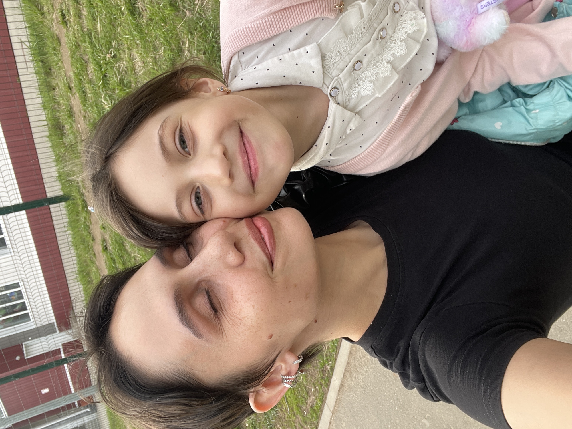 A selfie shows a woman with short dark hair, fair skin, and multiple earrings in her left ear, lying on her back with her eyes closed, while a young girl with long brown hair, fair skin, and a pink top leans on her chest, smiling at the camera. Behind them is a green grassy area and a building with a red and white exterior, which appears to be in Izhevsk, Russia. The girl is also wearing a white blouse with small black hearts, a pink cardigan, and holding a plush toy. The woman is wearing a black t-shirt. They are on a paved surface, possibly a sidewalk or road.